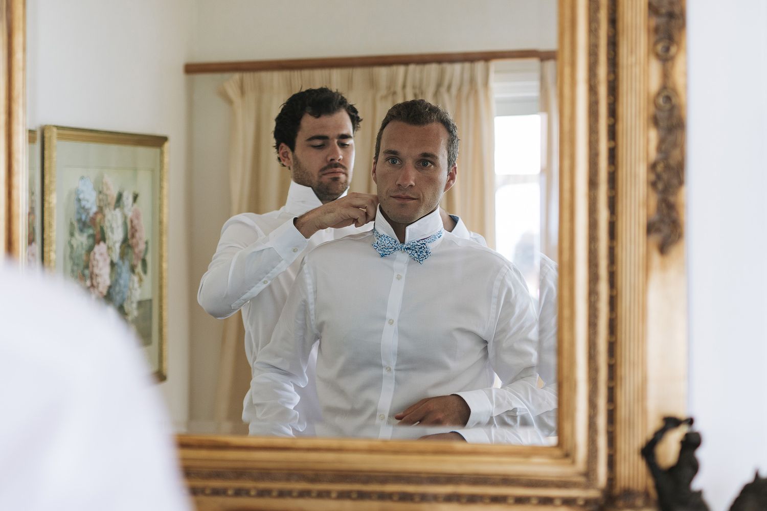 Two people in white dress shirts stand at a mirror getting ready for a formal occasion.