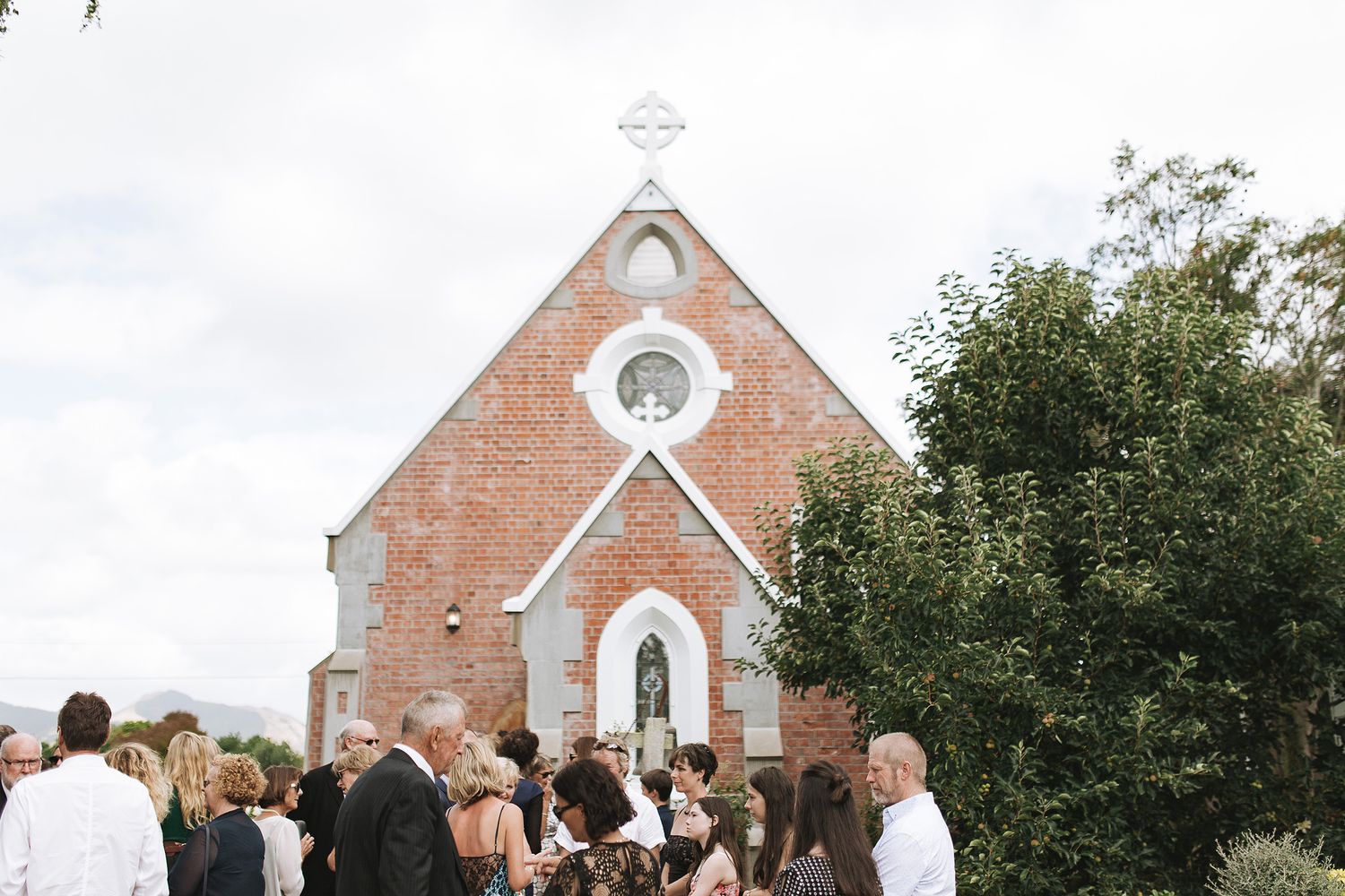 People gather outside a small brick church with arched windows and a cross on top for a special occasion.