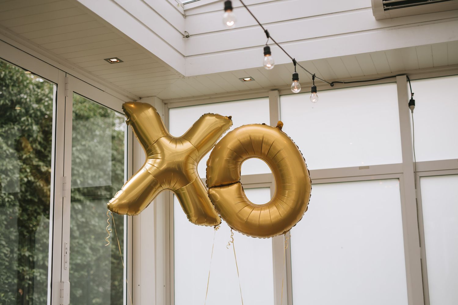 Gold metallic XO balloon letters floating in a bright sunlit room with white walls and string lights overhead.