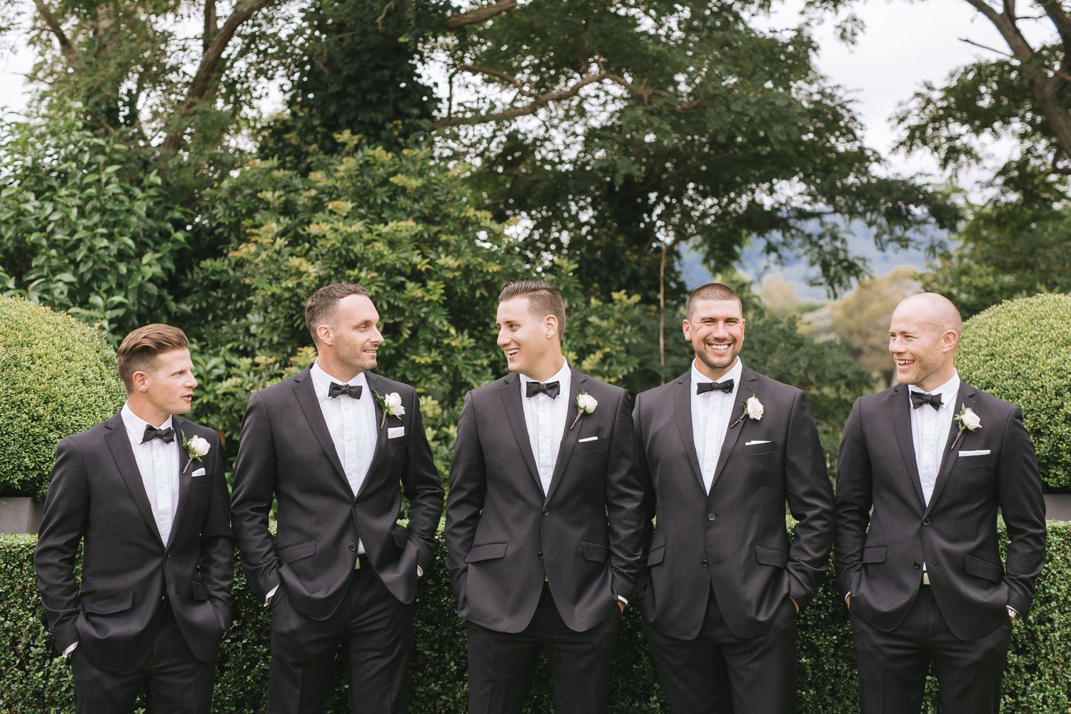 Groomsmen in black tuxedos standing in line outdoors sharing happy laughs in a garden setting.