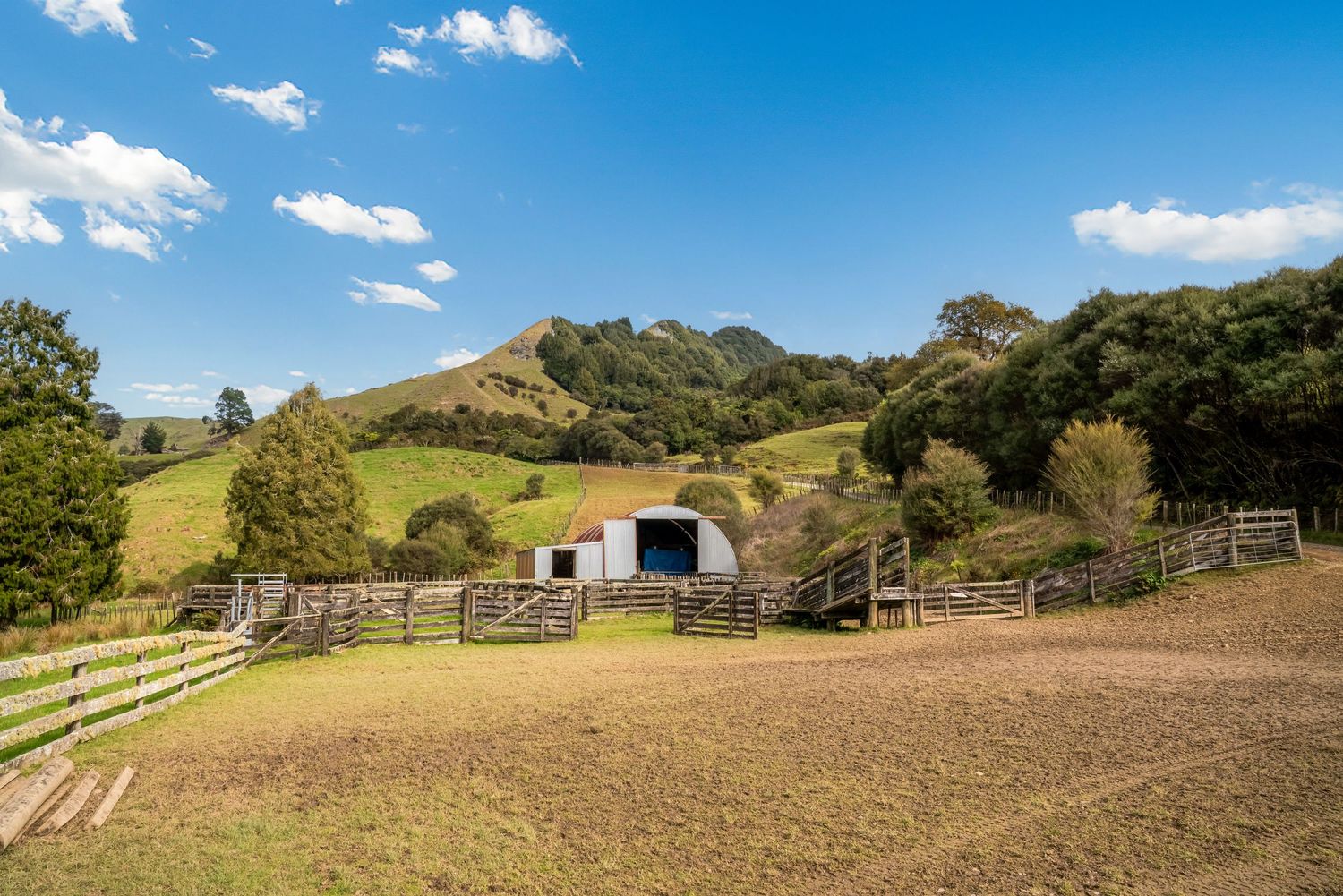 Rural barn shelter nestled in rolling green hills with wooden fencing and blue sky in New Zealand countryside.