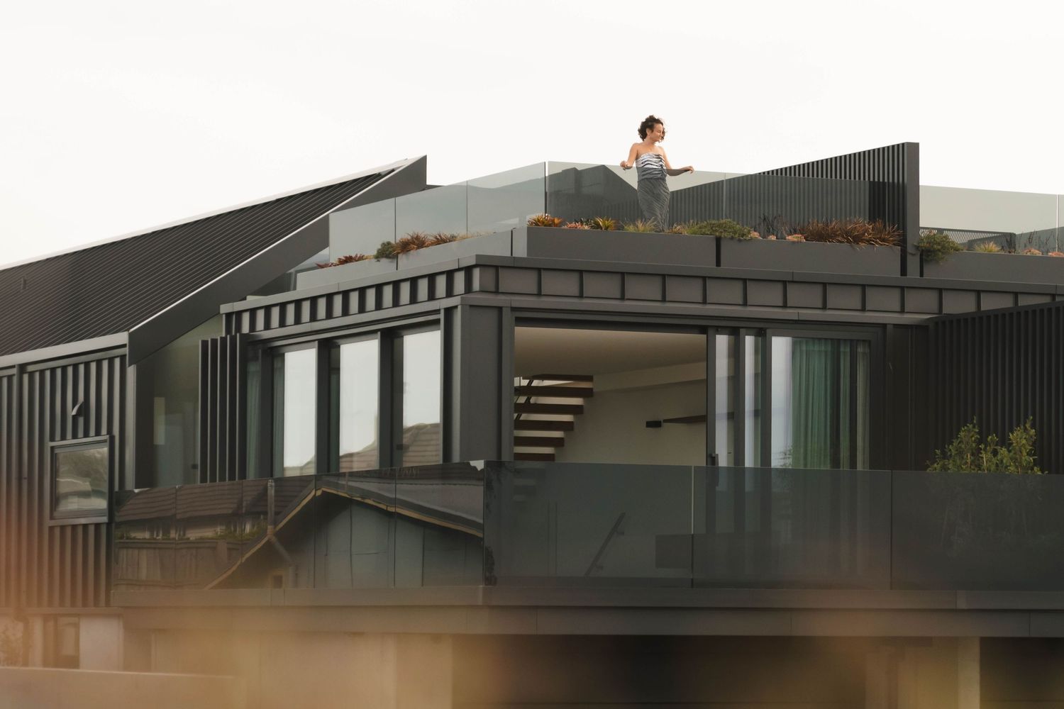 A person sits on the rooftop of a modern gray house with geometric architecture at dusk.