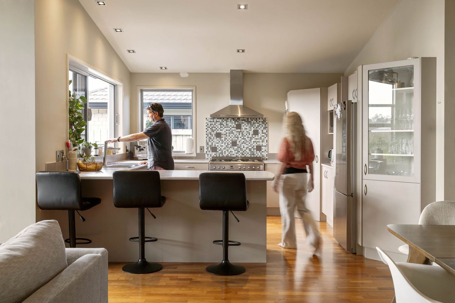 Modern open concept kitchen and living room with hardwood floors, black bar stools, and natural light from large windows.