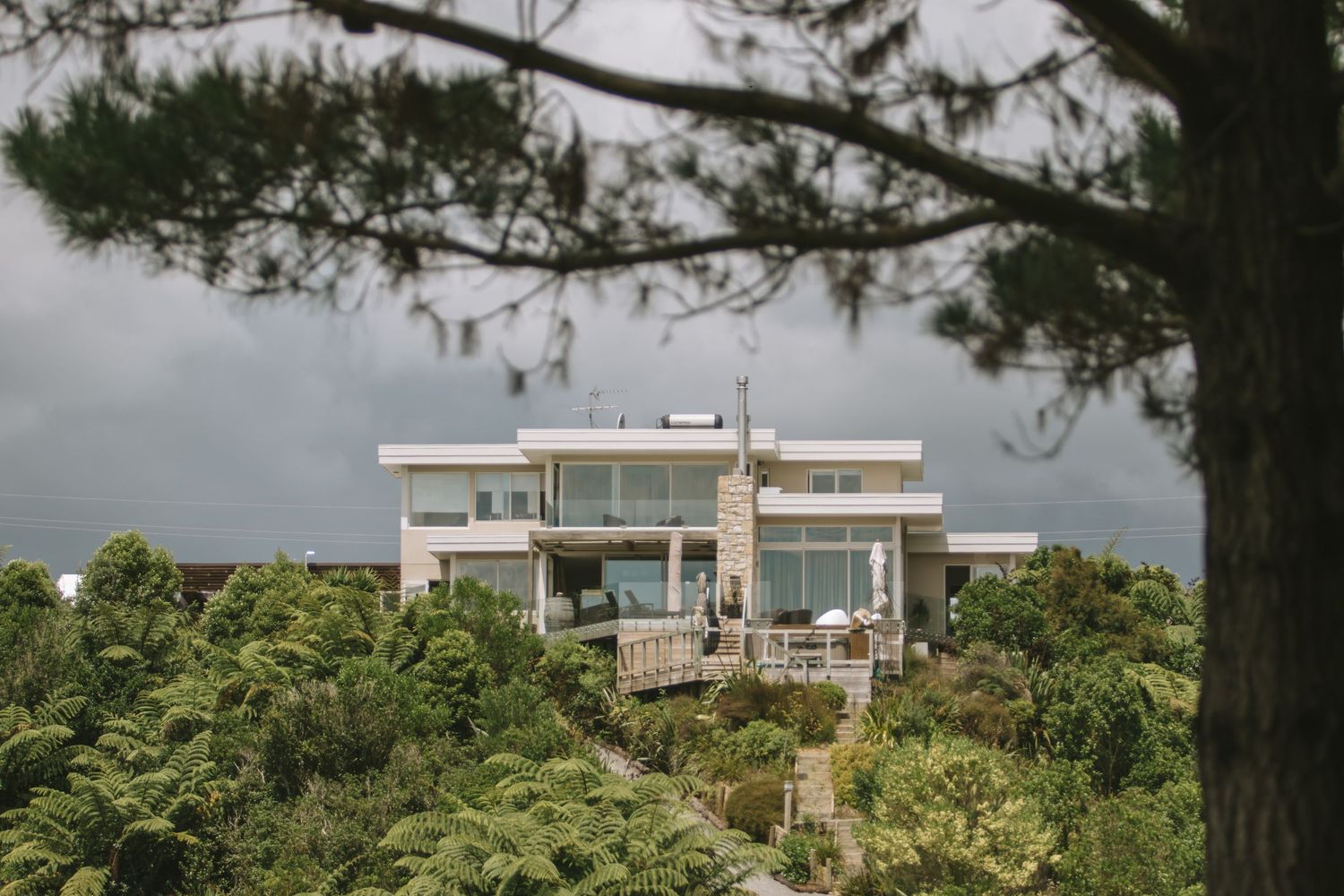 Modern luxury hillside house with floor-to-ceiling windows nestled among lush green trees against a moody sky.