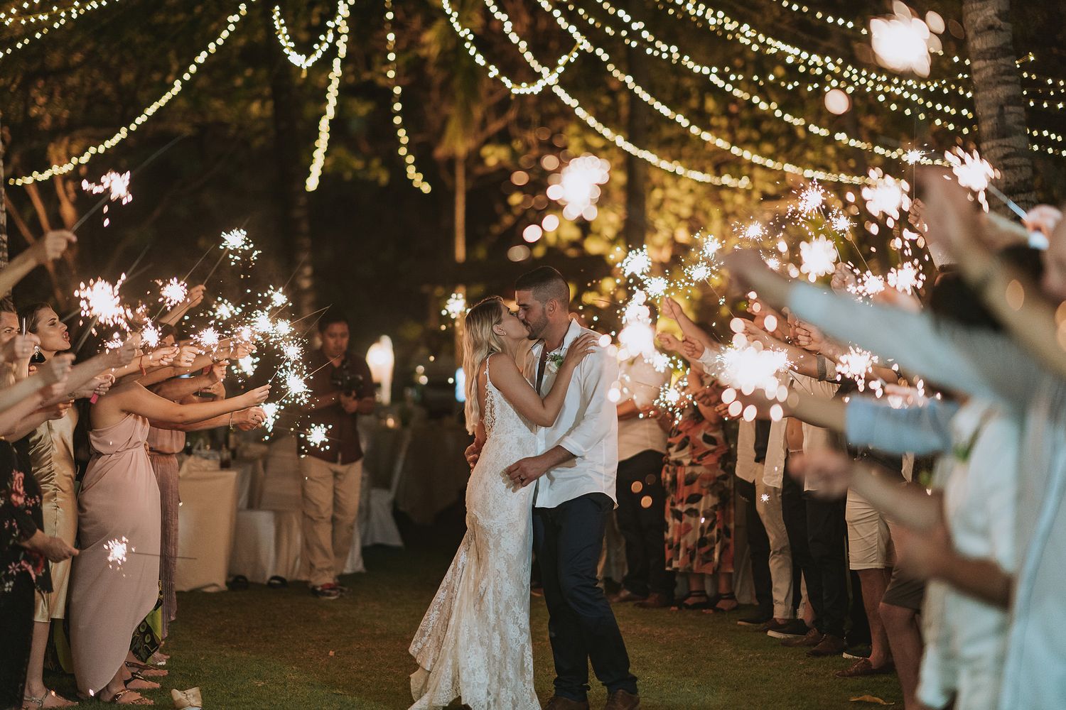 Couple shares romantic first dance outdoors under twinkling string lights as guests hold sparklers at evening wedding reception.