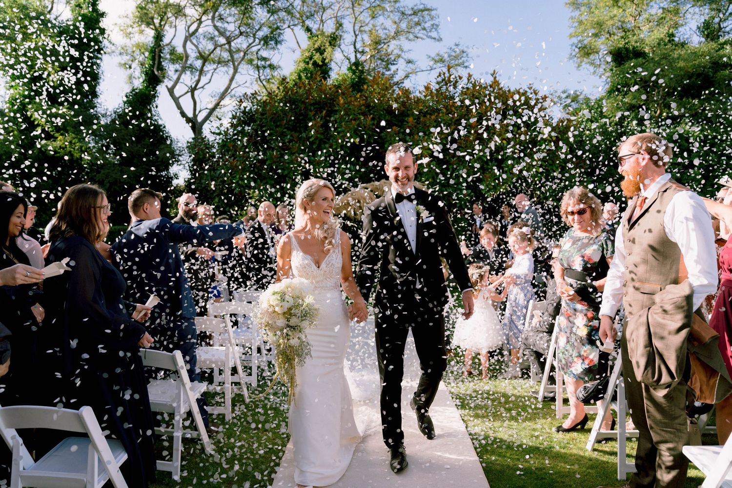Wedding couple walks down aisle while guests throw confetti in celebration at outdoor garden ceremony.