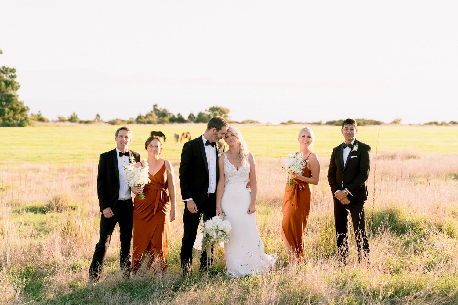 A wedding party standing in a golden field at sunset wearing rust-colored and black formal attire.