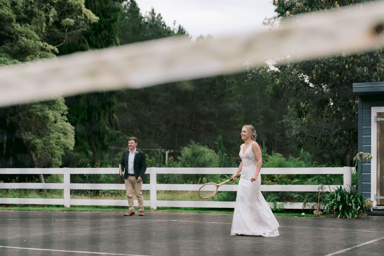 A couple stands apart on a wet deck surrounded by white fencing and green trees.