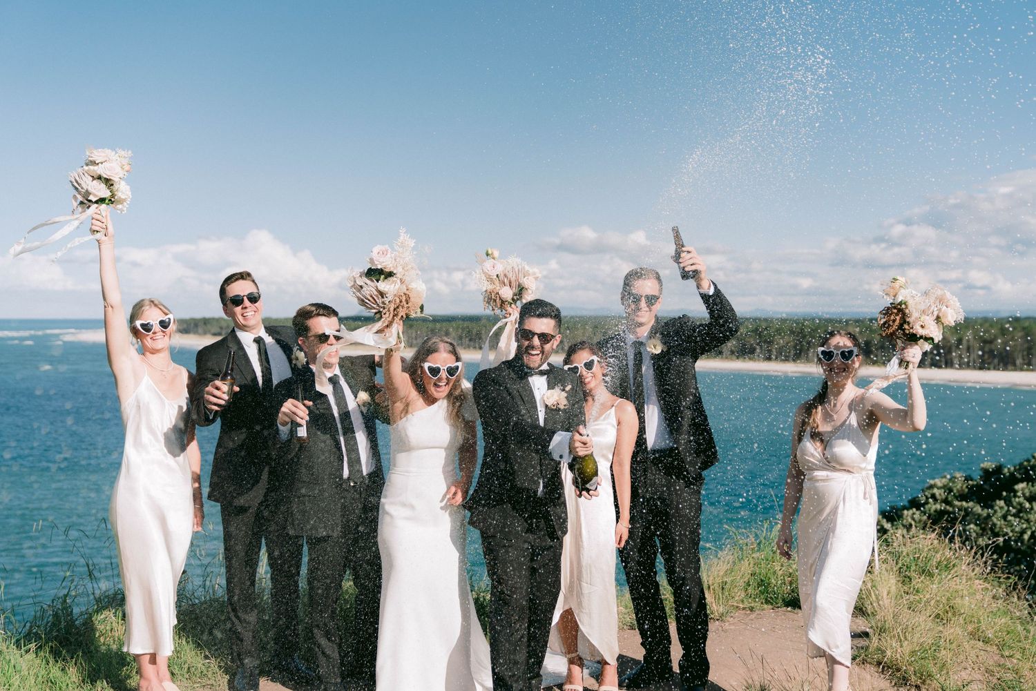 Wedding party celebrates joyfully against an oceanfront backdrop.