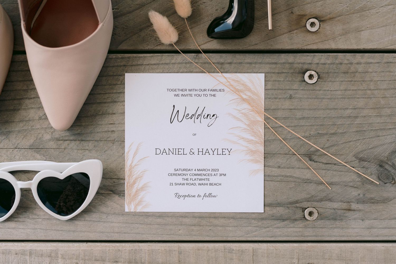 Wedding invitation with pink details lies on wooden surface alongside heart-shaped sunglasses and pale pink shoes.