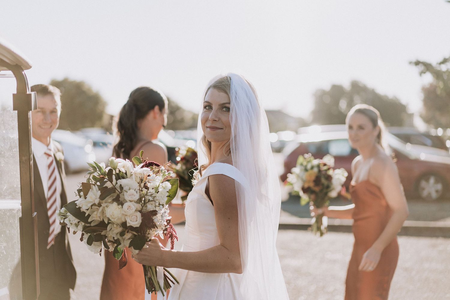 Bridal party gathers outdoors in warm sunlight holding elegant bouquets of white and blush flowers.