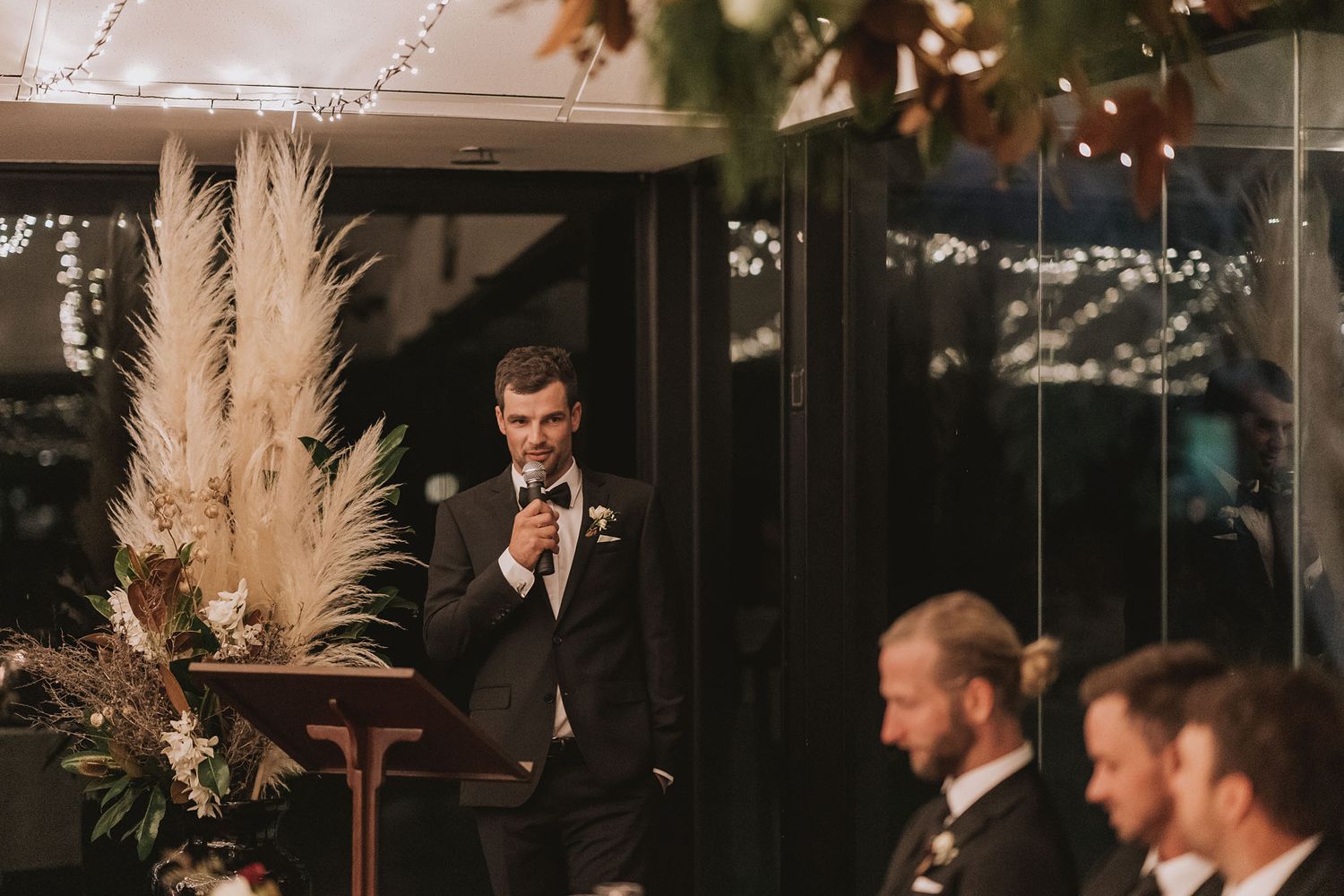 An emotional wedding speech being delivered at an evening reception decorated with pampas grass and string lights.