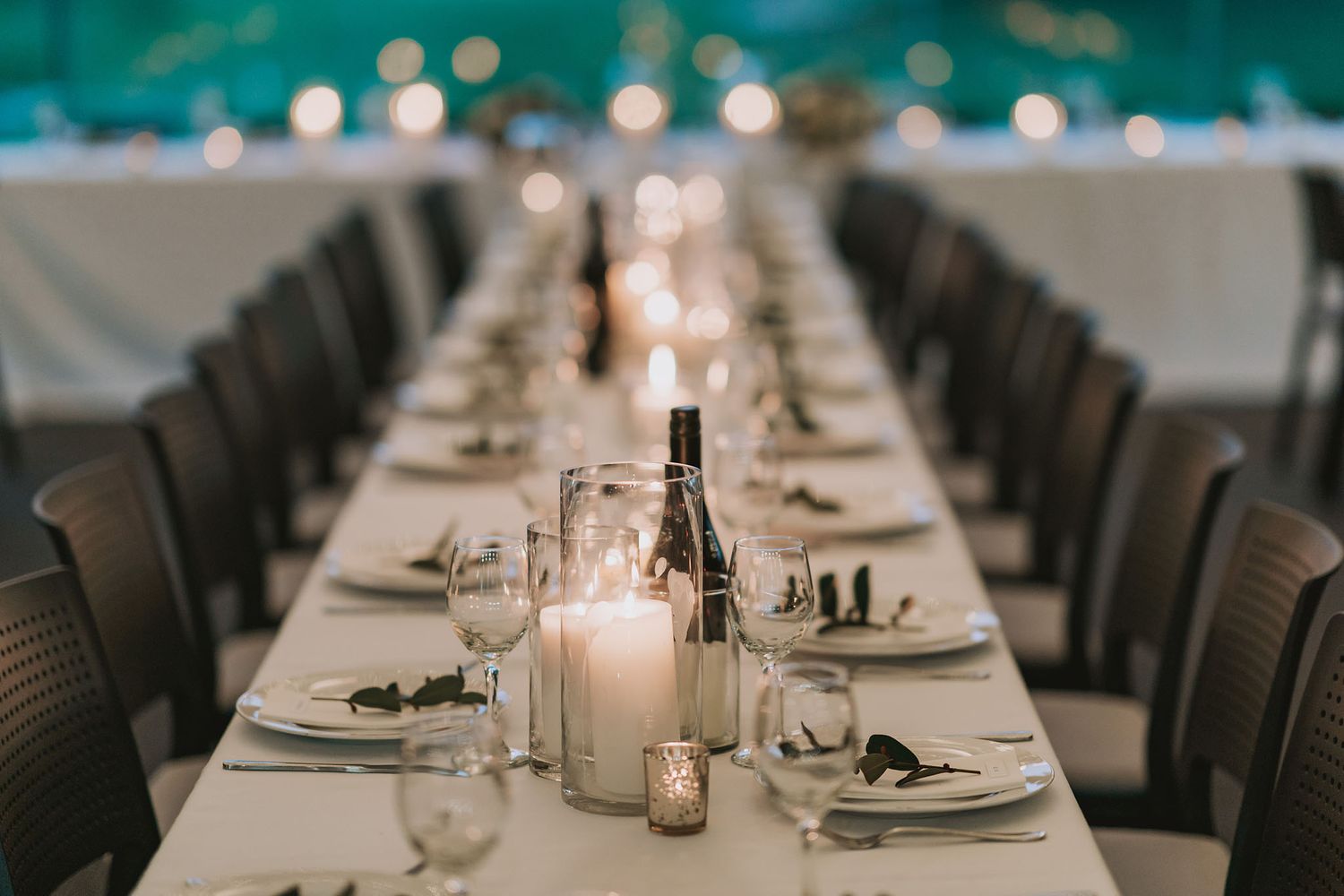 Long elegant dining table set for a wedding reception with candles and place settings in soft ambient lighting.