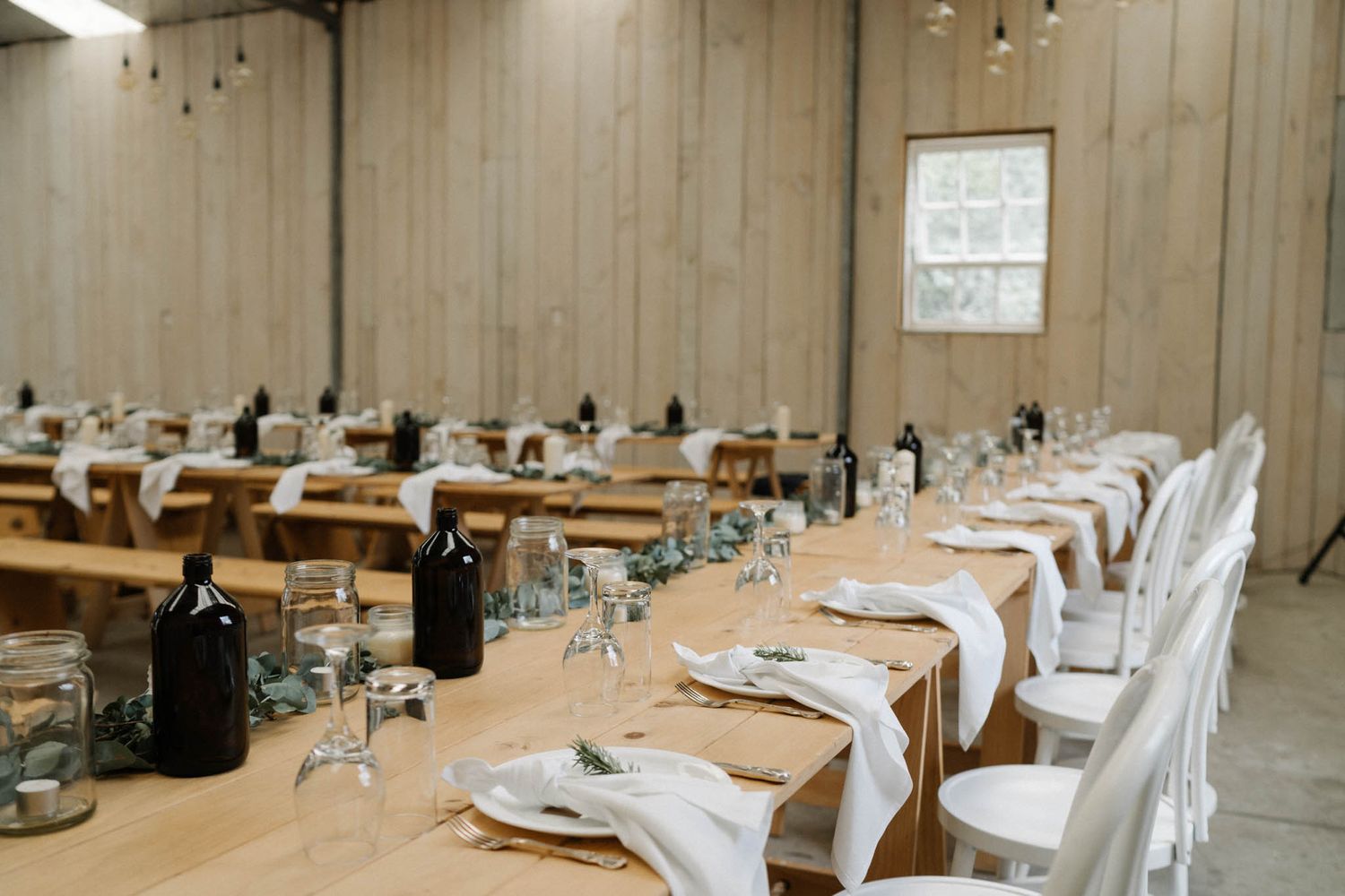Long banquet tables set with elegant white linens and chairs arranged in a barn reception hall for a wedding celebration.