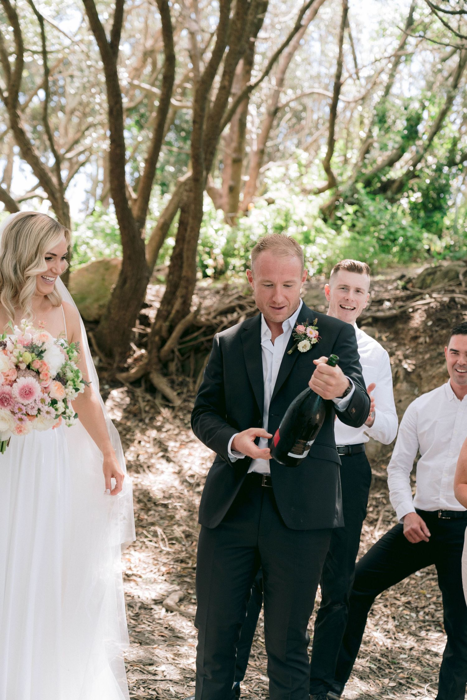 Wedding party celebrates in an outdoor wooded setting with candid reactions and natural lighting.