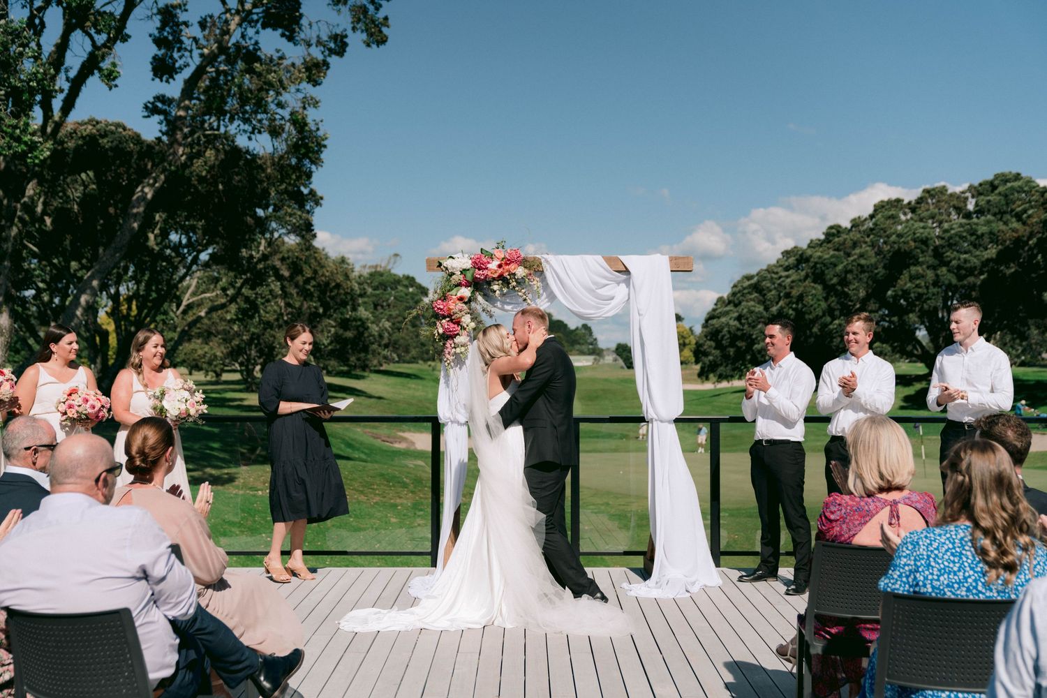 Wedding ceremony on outdoor deck with couple exchanging vows under white arch with guests seated.