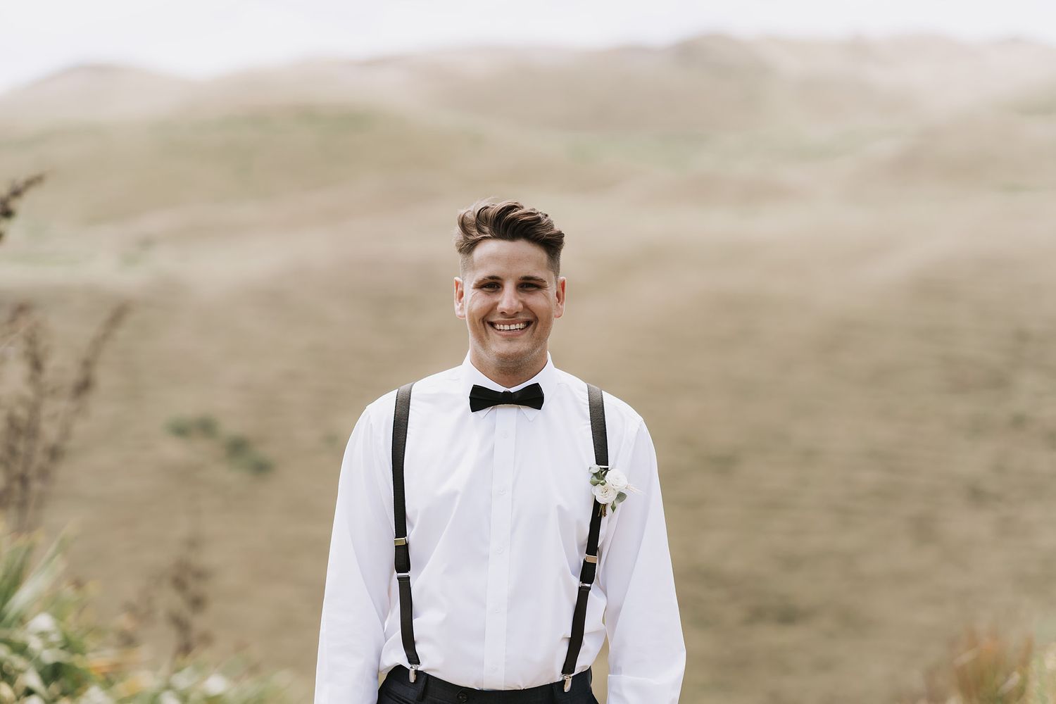 A person in a white shirt and black suspenders stands smiling against a blurred outdoor background.