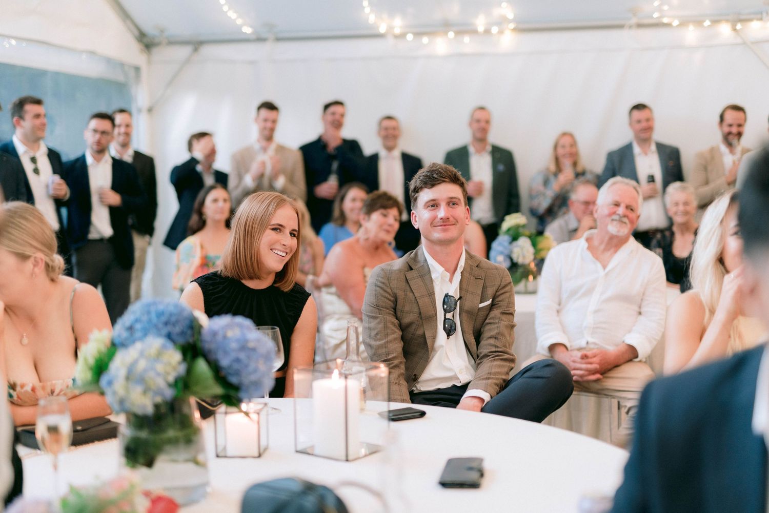 Wedding guests seated at round tables under a white tent with blue hydrangea centerpieces and string lights.