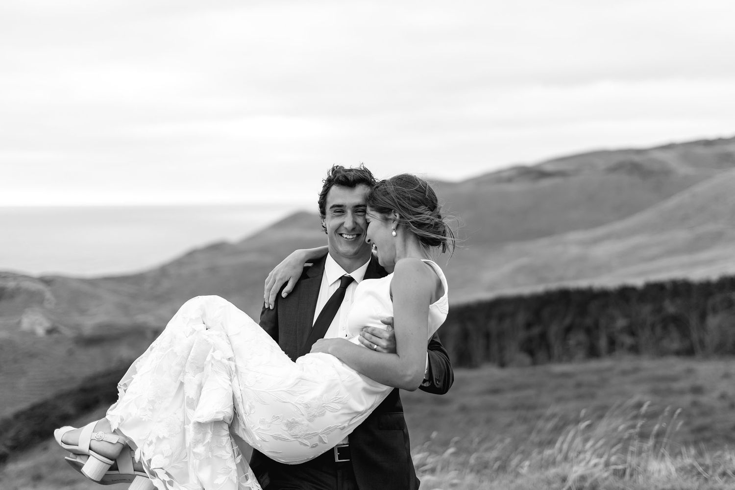 Bride and groom share romantic moment outdoors with sweeping mountain landscape in black and white.