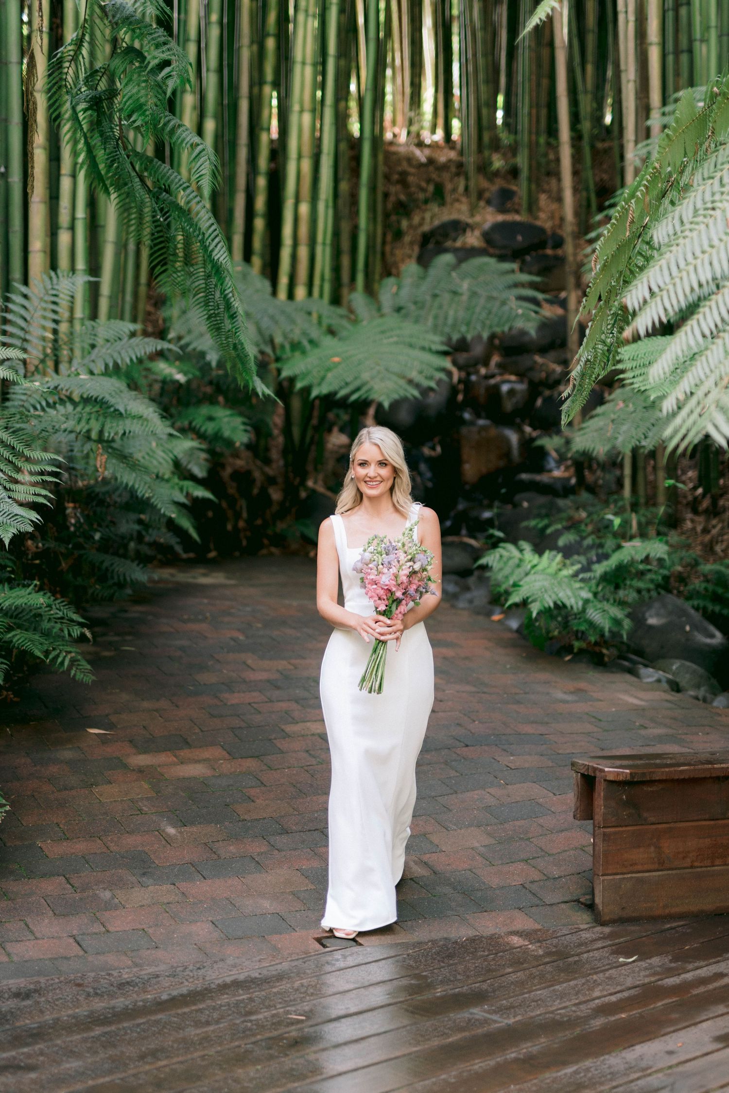 A bride in a white wedding gown walks through a bamboo garden pathway holding a colorful bouquet.