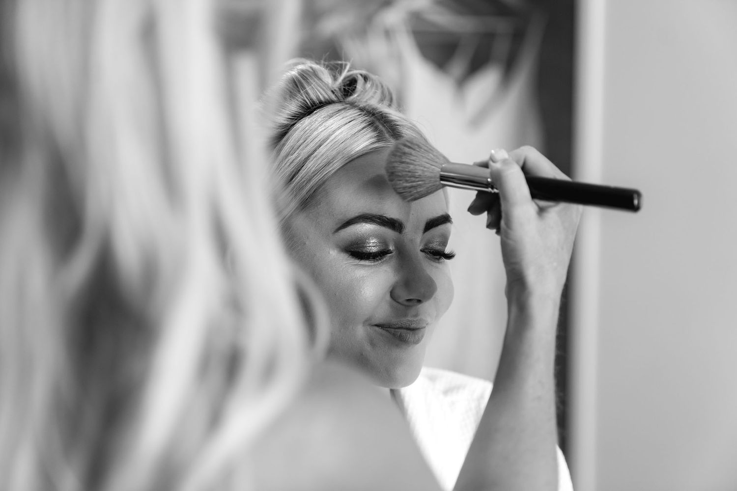 A black and white close-up of someone having makeup applied with a brush near their eye.