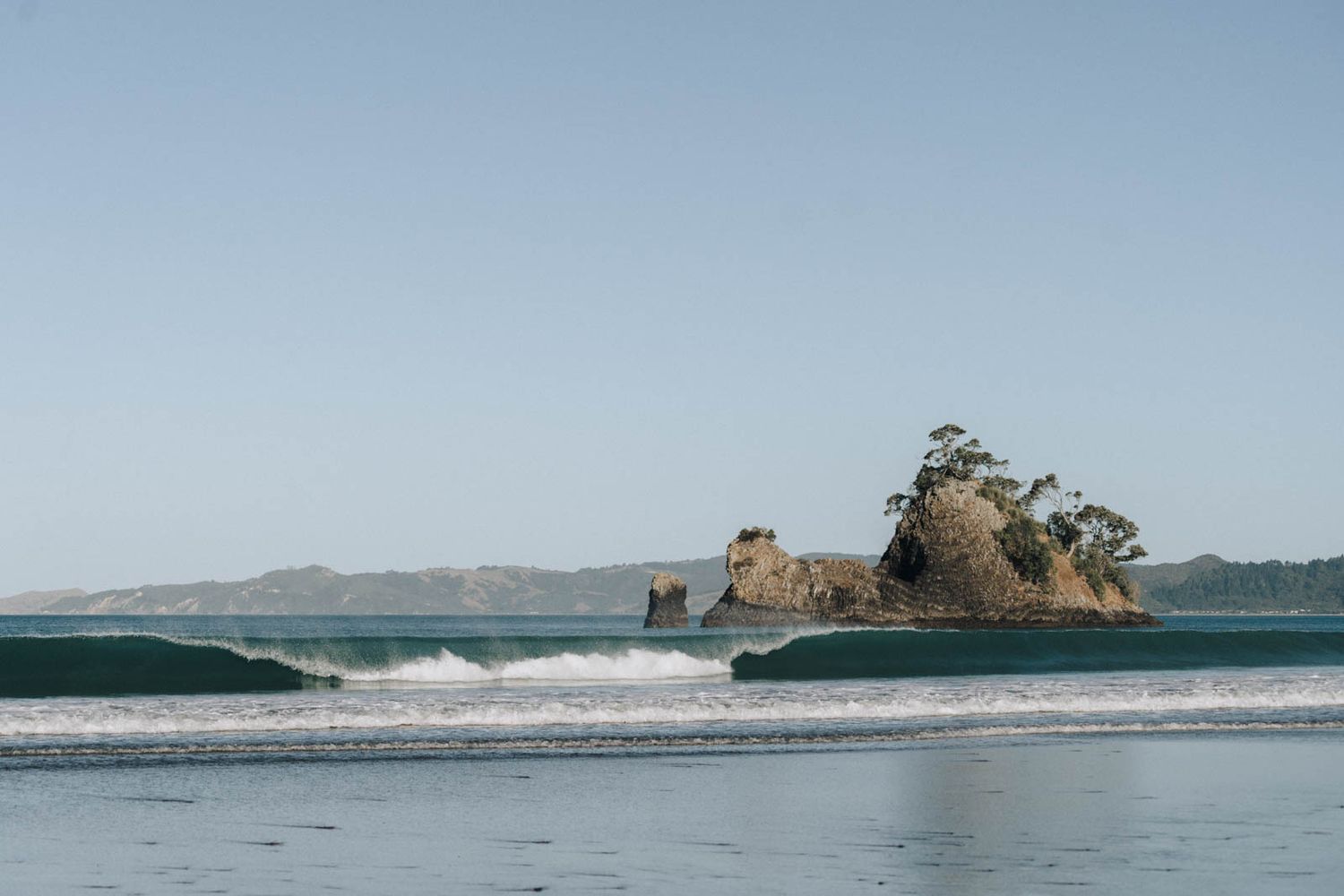 Waves crash along a scenic coastal shoreline with a rugged rocky outcrop jutting into the tranquil ocean waters.