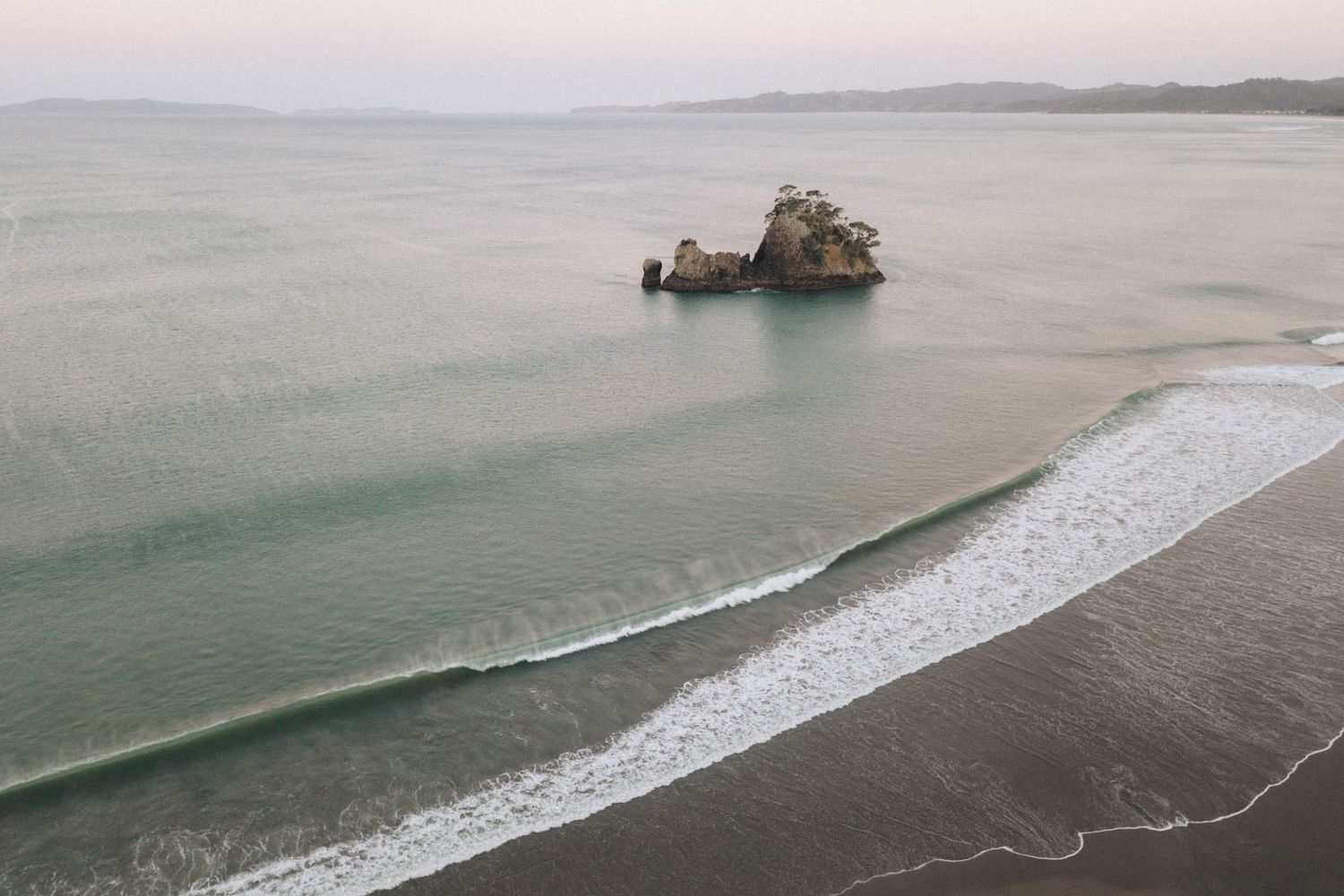 Aerial view of a dramatic coastline with a lone rock formation in the calm ocean and gentle waves rolling onto shore.