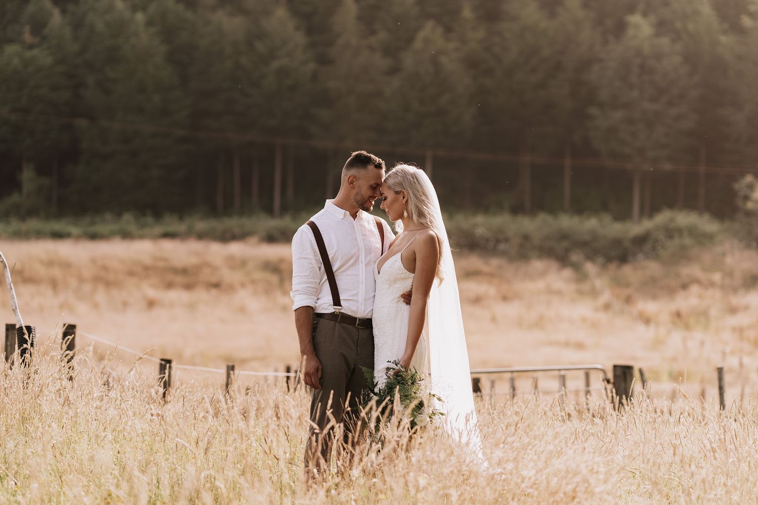 A romantic couple in wedding attire embraces while standing in a rustic field during sunset hour.