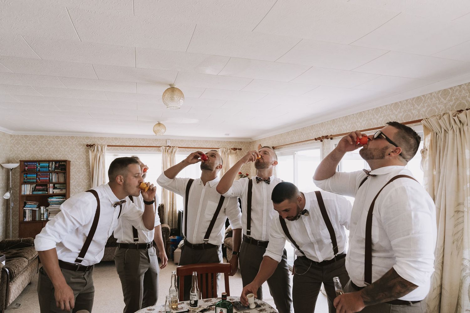 Groomsmen wearing white shirts and suspenders pose together while getting ready in a bright hotel room.