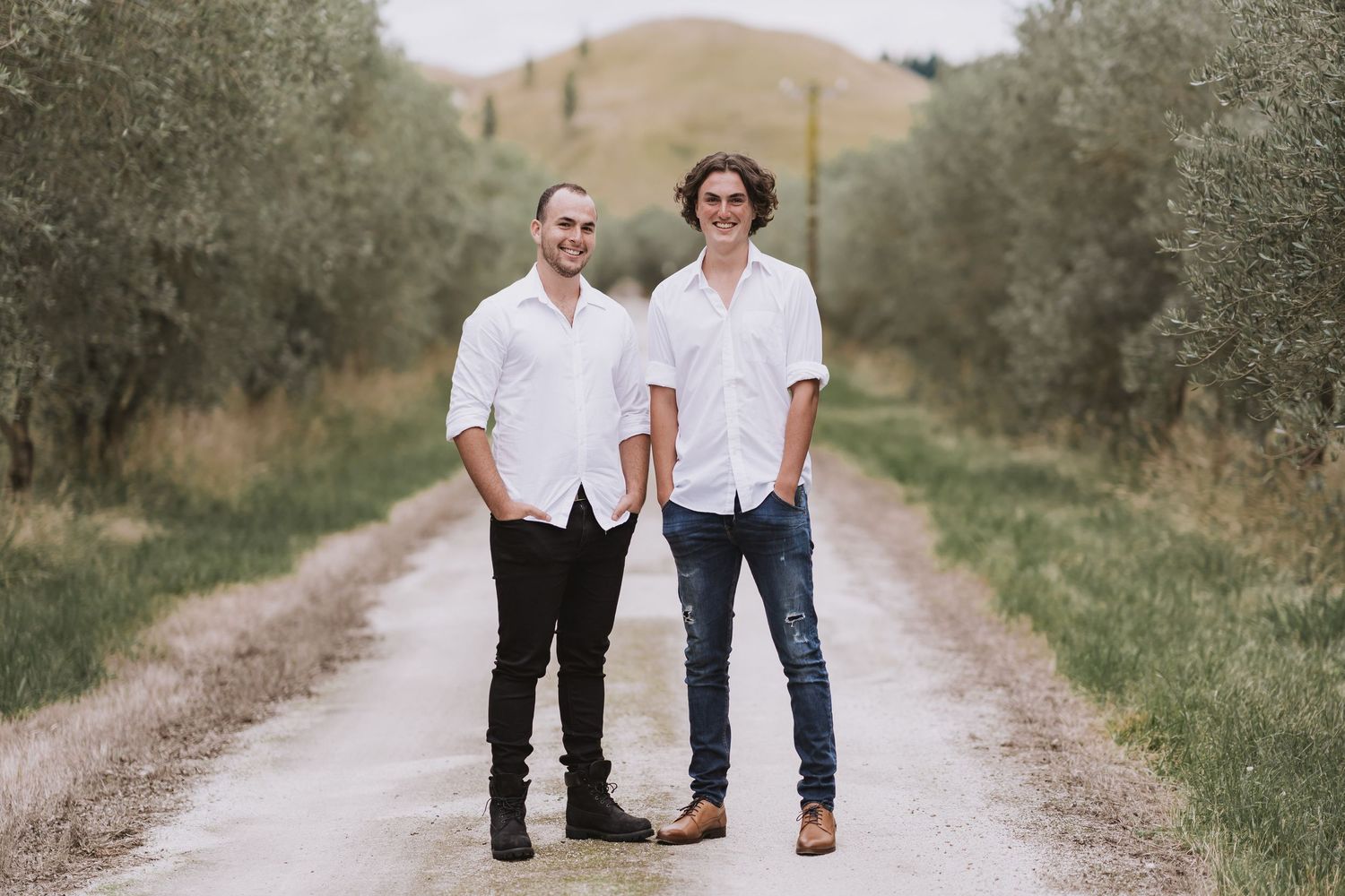 Two people in matching white shirts and dark pants walk together on a rustic dirt path surrounded by olive trees.