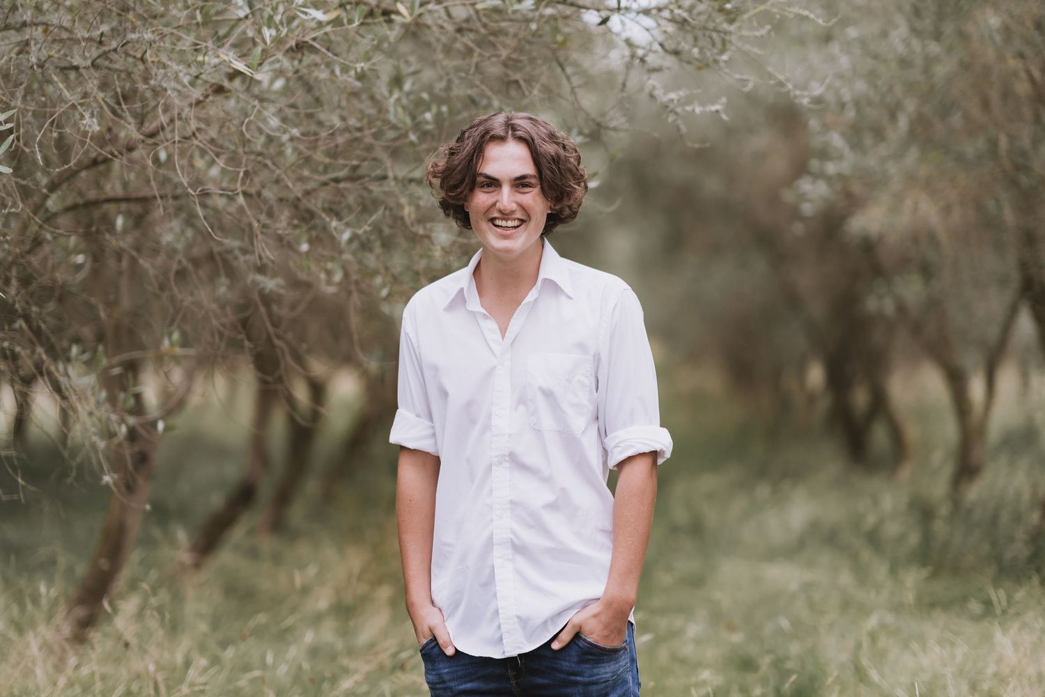 A person in casual white button-down shirt smiling while standing against a blurred natural background.