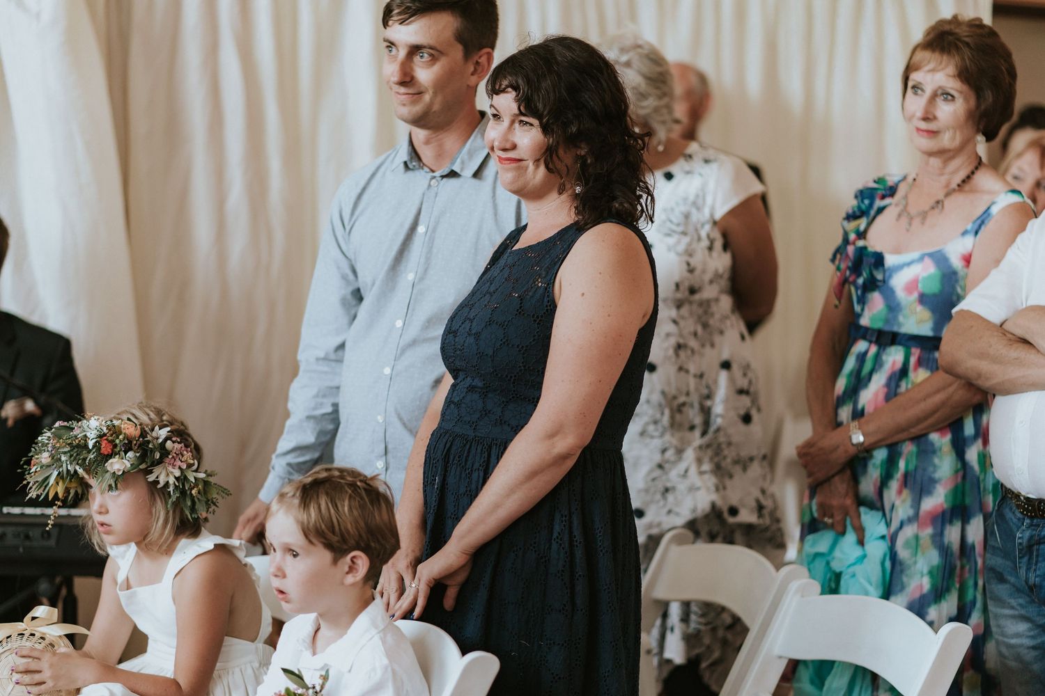 Wedding guests stand together watching a celebration in a room with white curtains as backdrop.