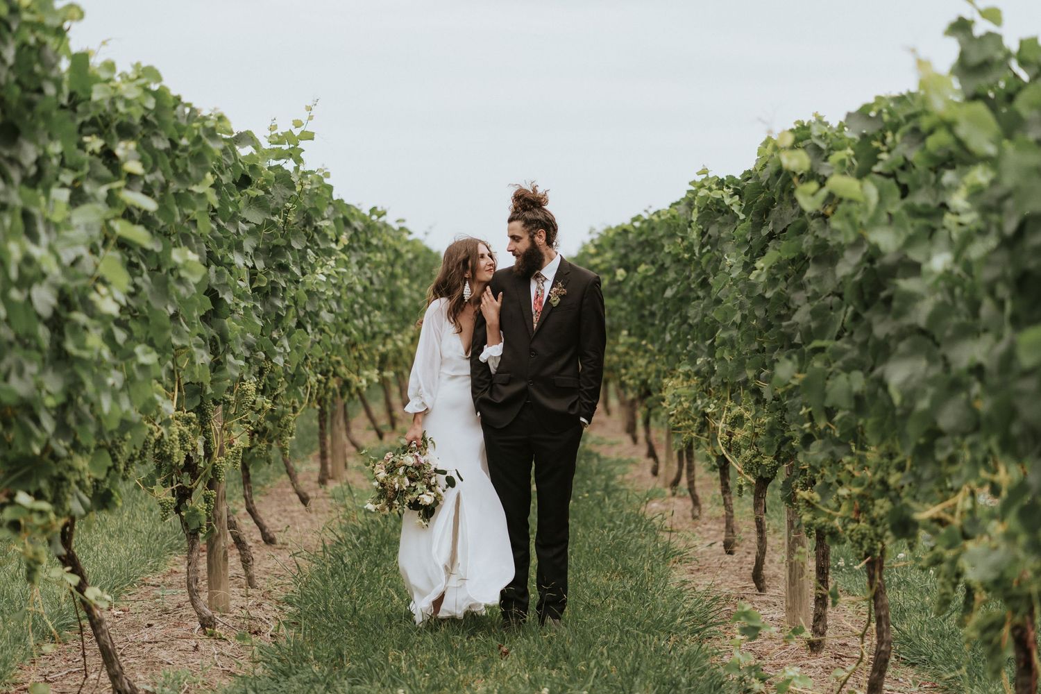 A couple stands intimately together between rows of grape vines at a scenic vineyard wedding.
