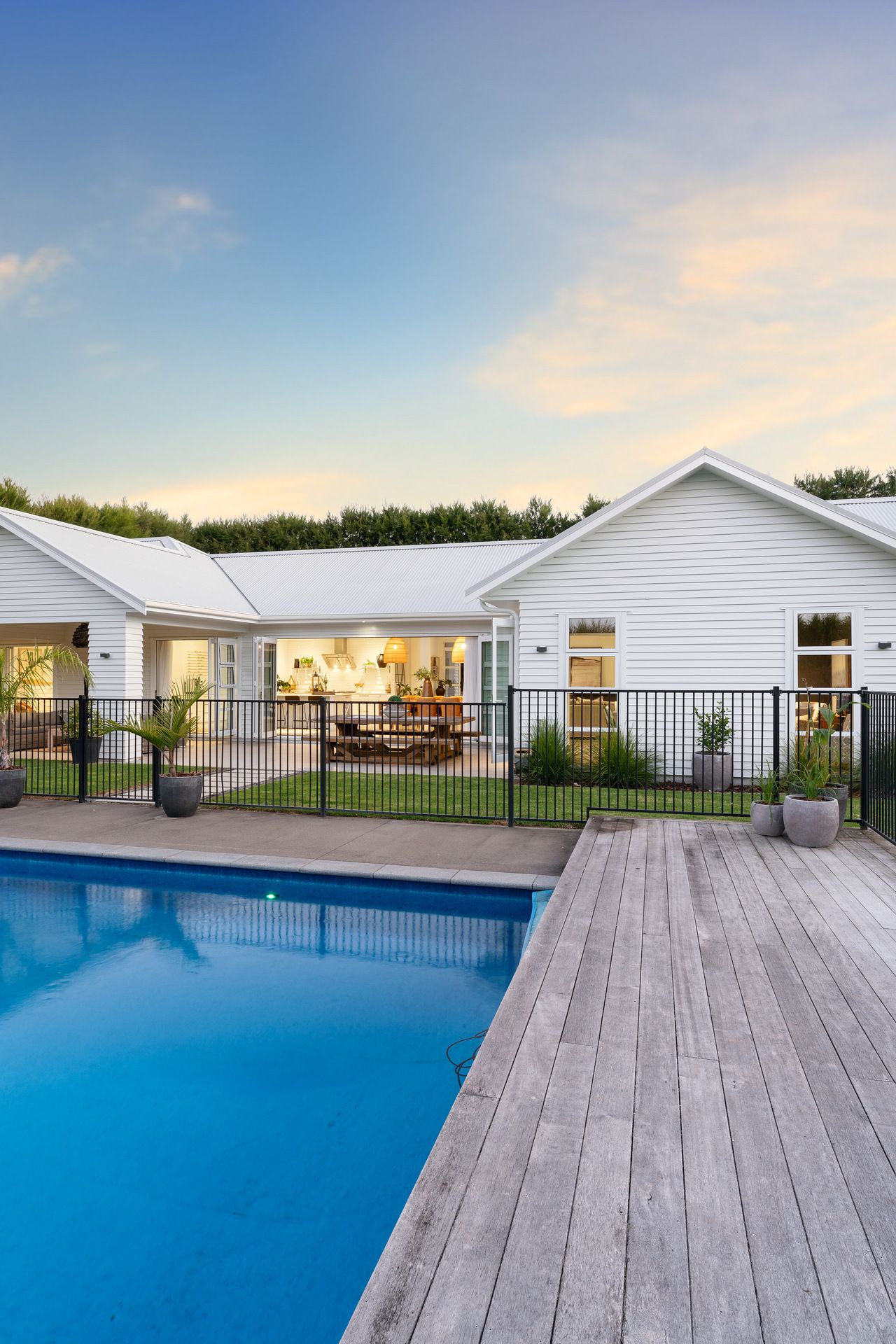 Modern white house with wooden pool deck and illuminated interior visible through glass doors at dusk.