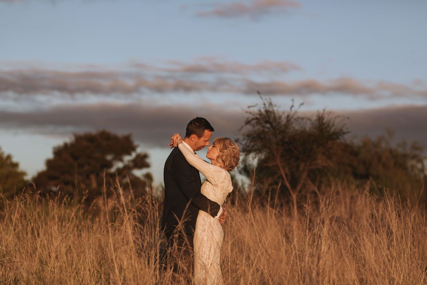 A romantic couple embraces in a golden field at sunset with clouds and trees in the background.