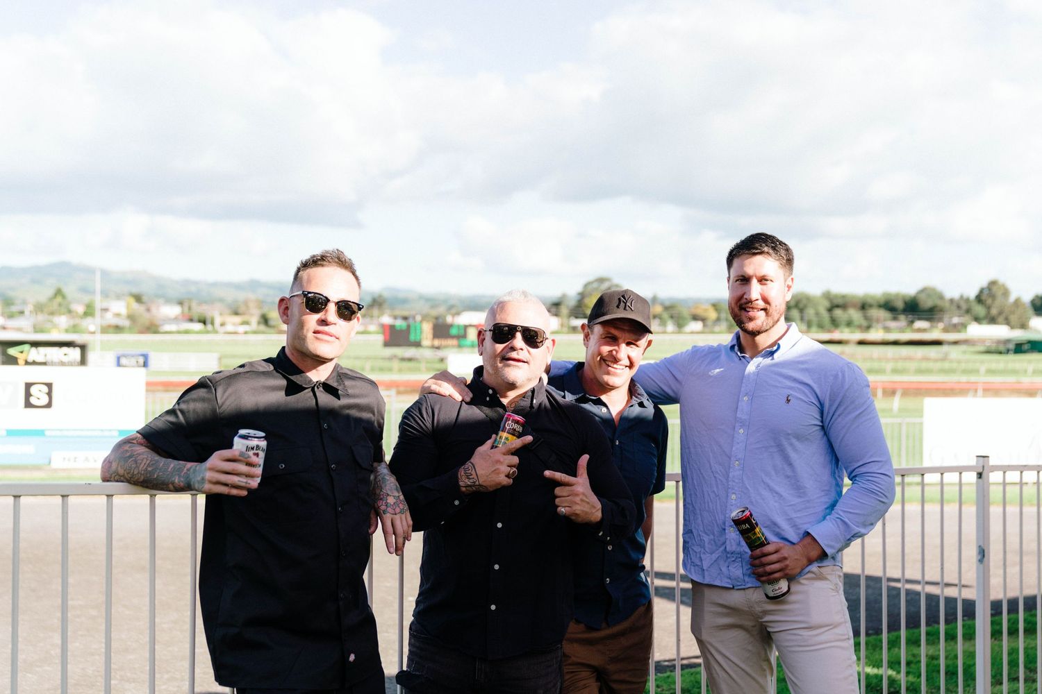 Four friends holding drinks pose together against a racetrack backdrop.