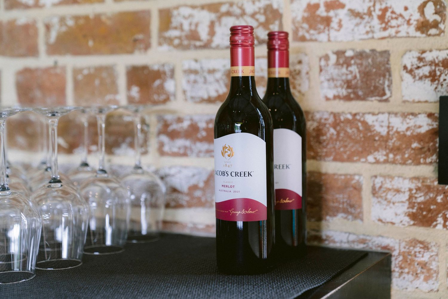 Two red wine bottles and empty wine glasses sit on a dark surface against an exposed brick wall.