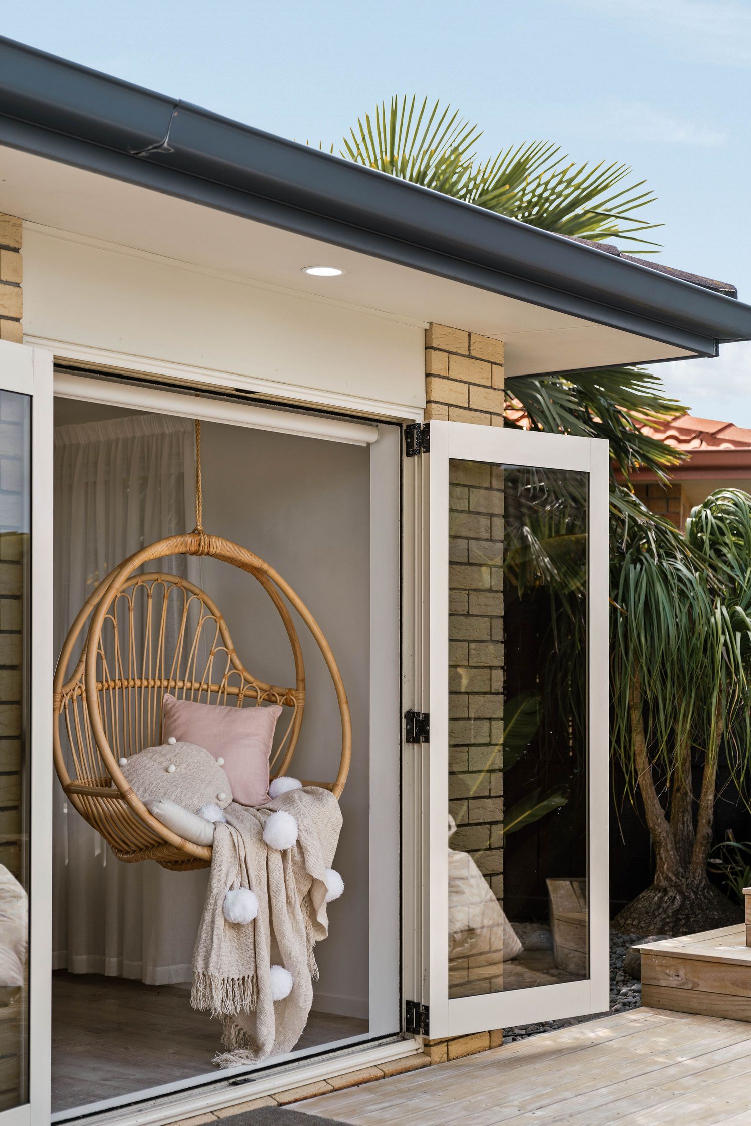 A hanging rattan egg chair with pink cushions and throw blankets sits by glass sliding doors with tropical plants outside.