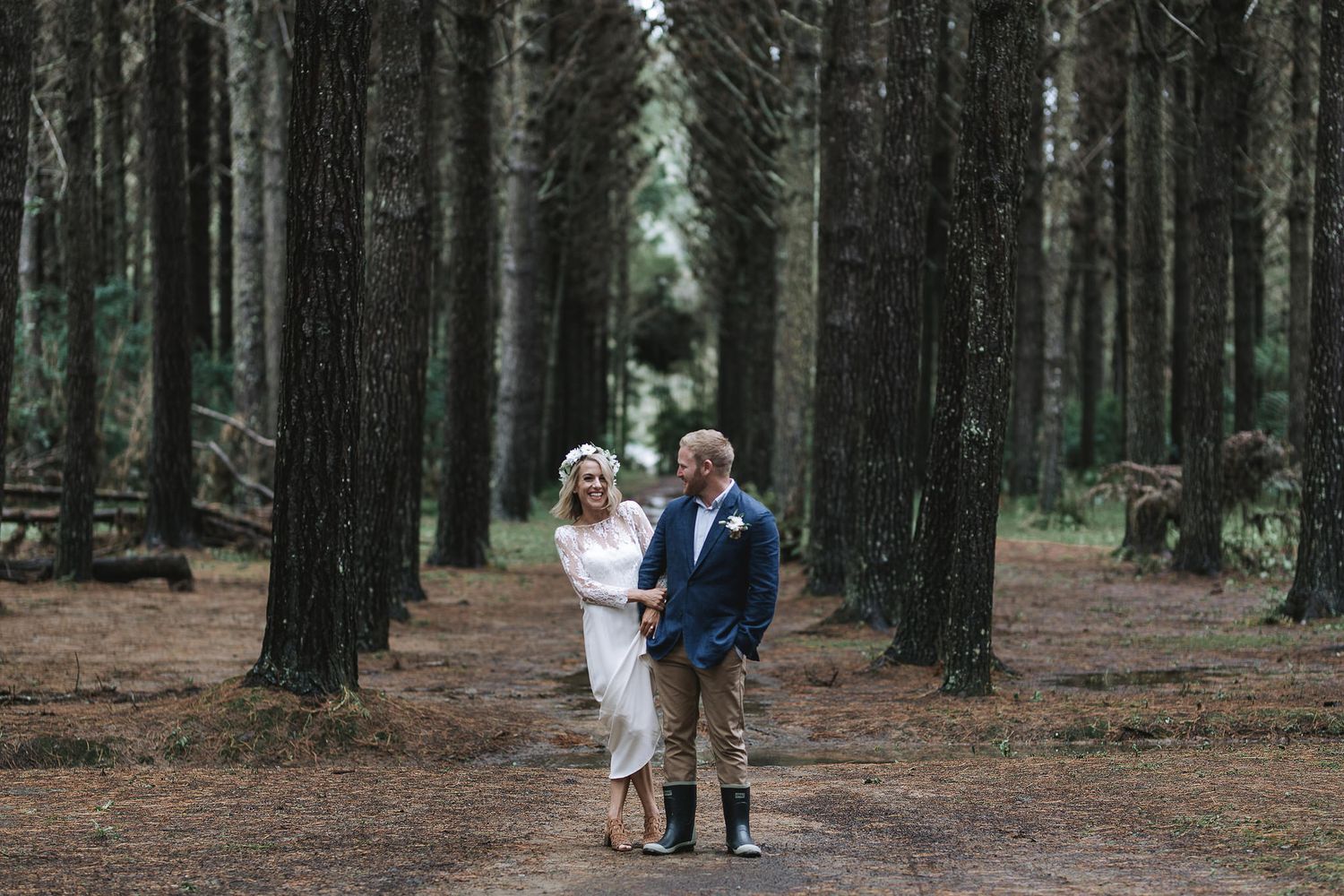 A couple stands together in a majestic evergreen forest with tall trees creating a symmetrical path behind them.