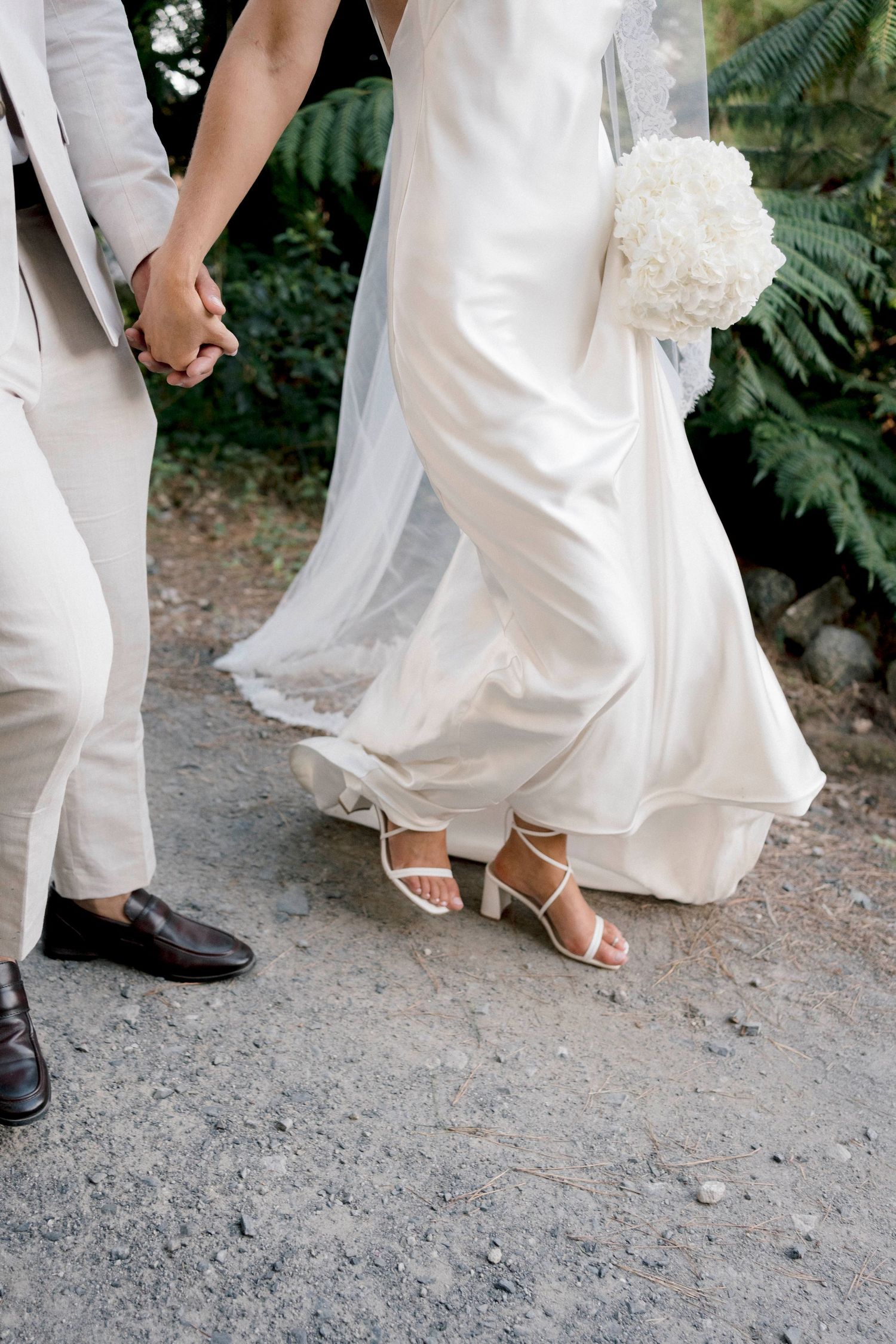 Close-up of bride and groom's feet and dress hem while holding hands on a gravel path.