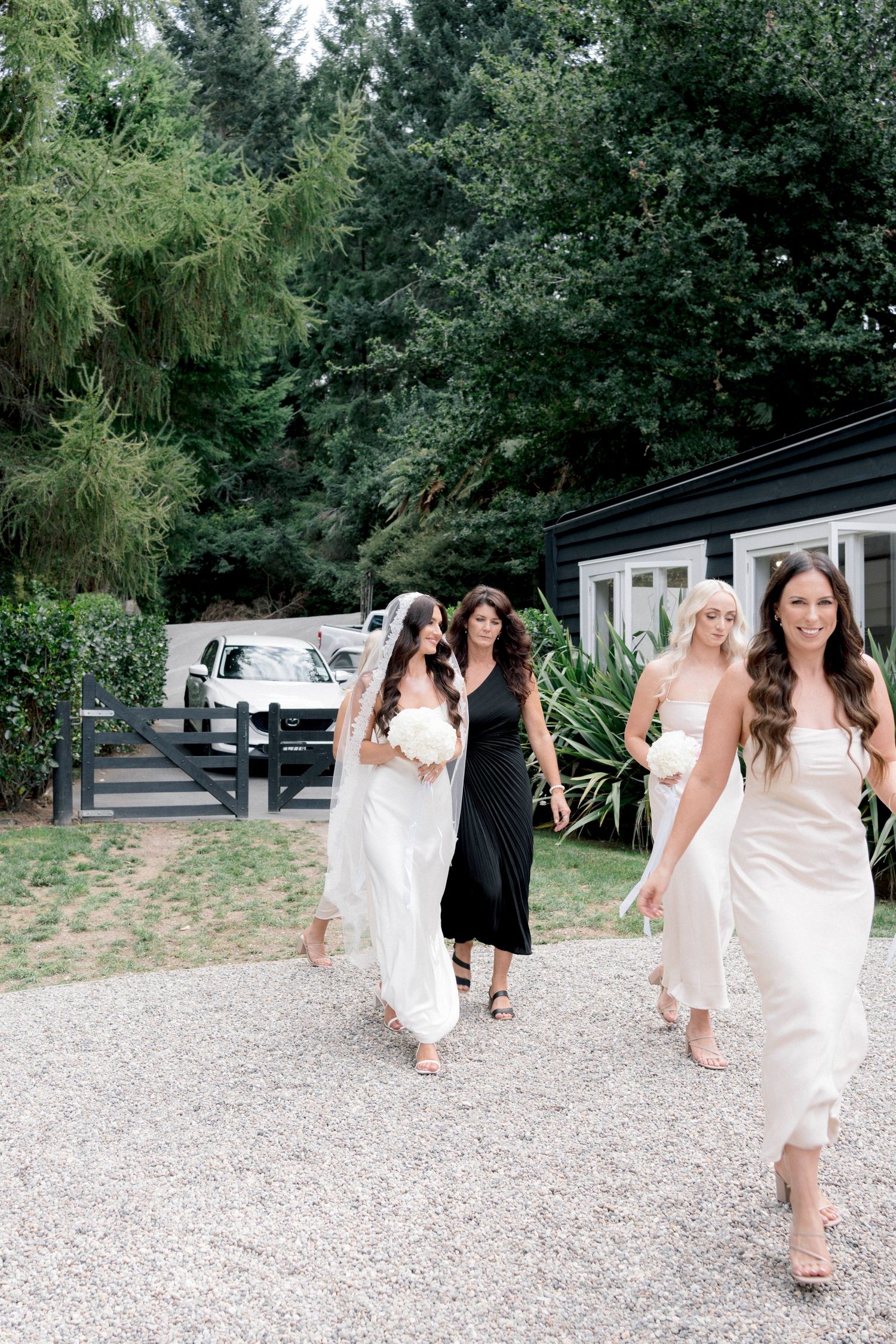 Bridesmaids in white dresses and one in black walking on gravel path near modern building.