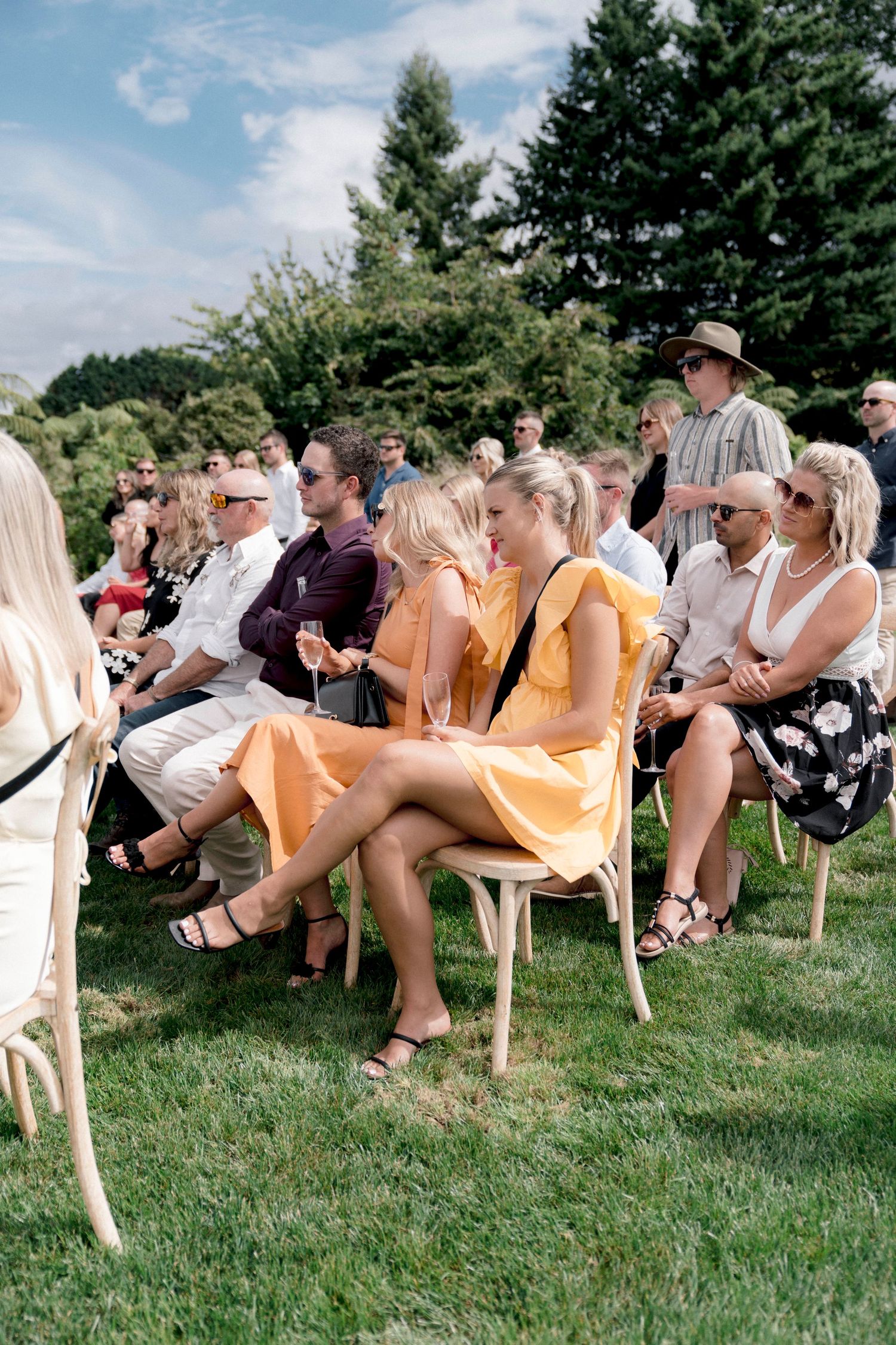 Wedding guests seated in wooden chairs on a lawn during an outdoor summer ceremony.