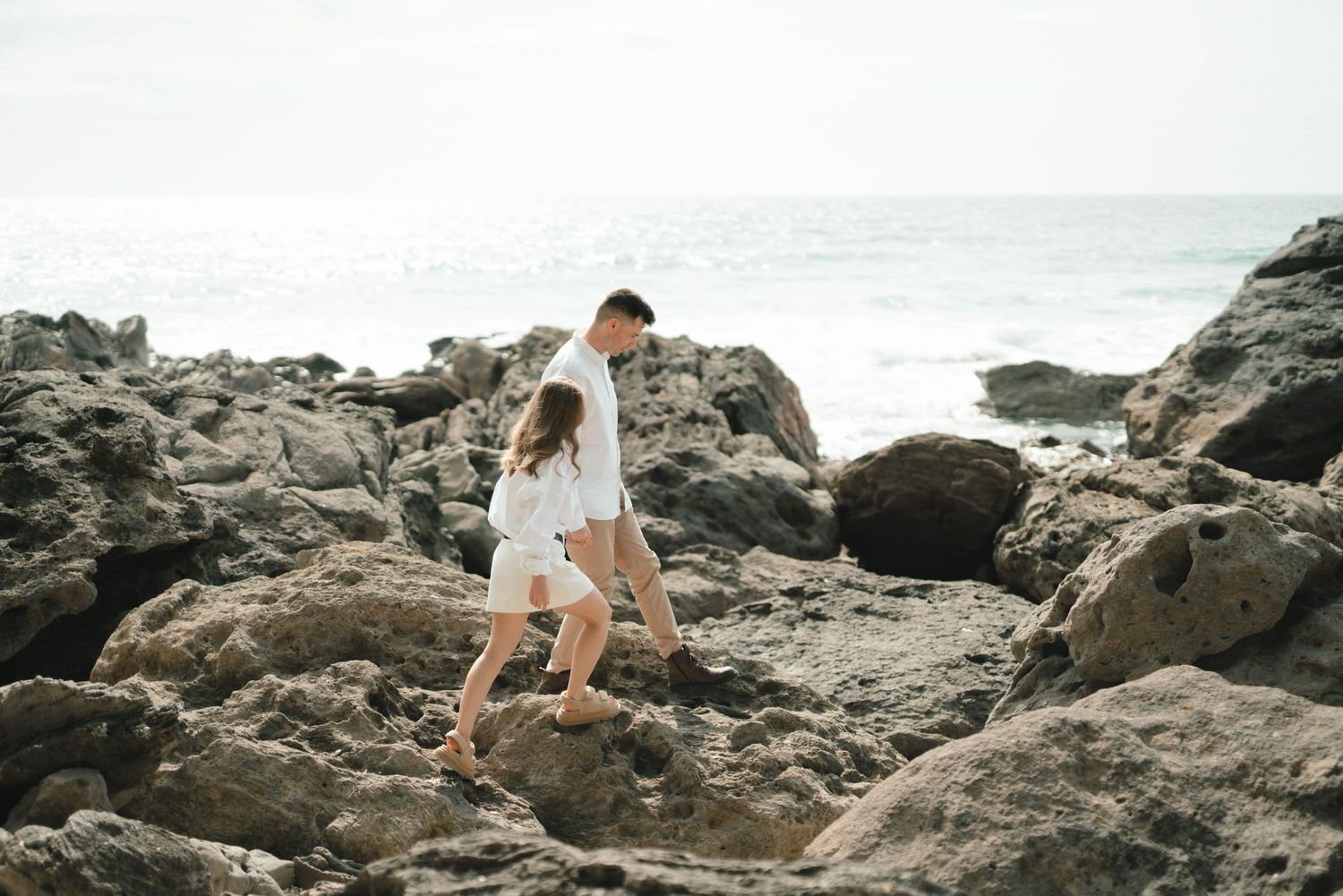 Two people in white outfits carefully navigate across rocky coastal terrain with ocean waves visible in the background.