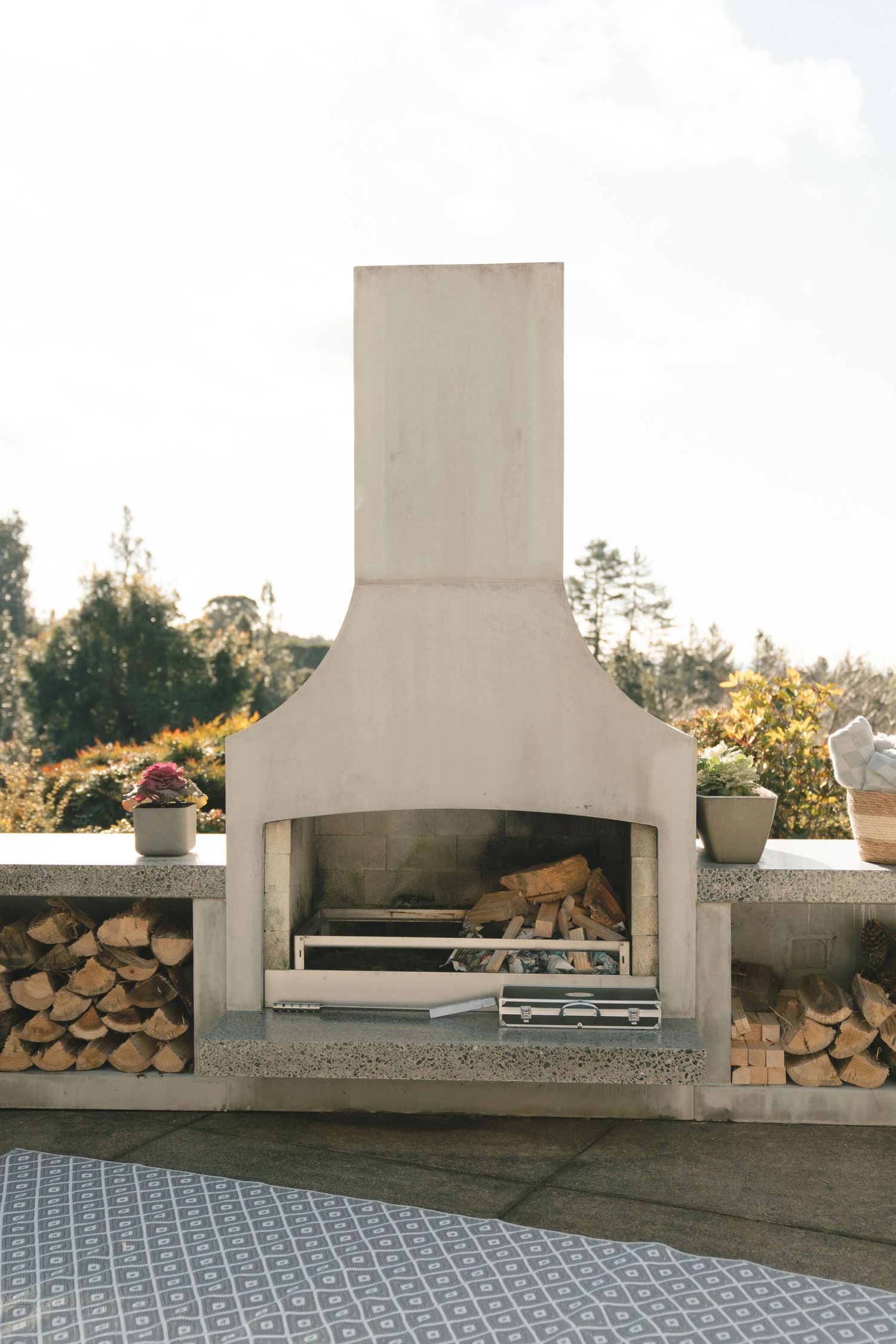 Modern outdoor concrete fireplace with white hood and built-in wood storage on a patterned tile patio.