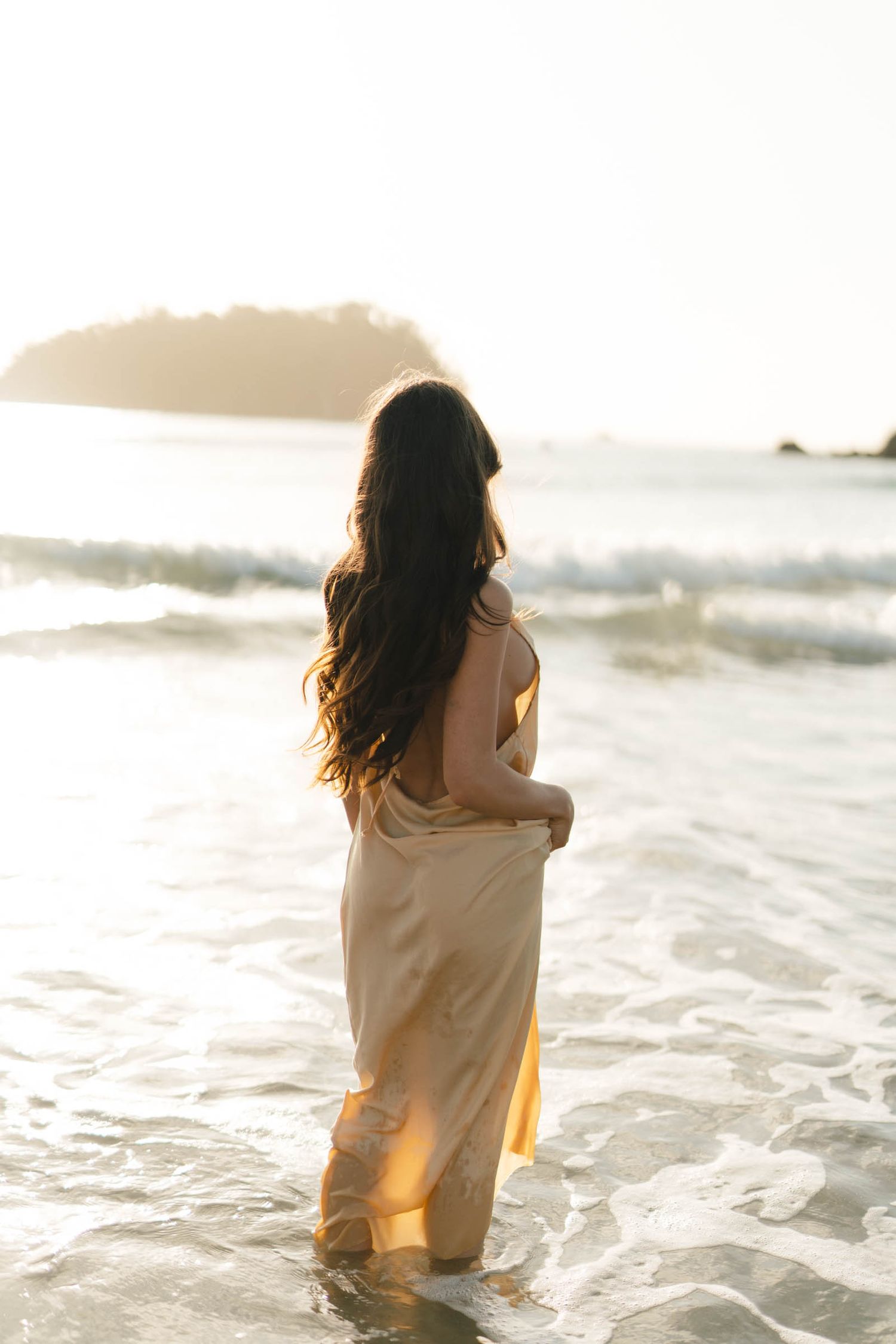 Person in light clothing stands in ocean waves during golden hour.