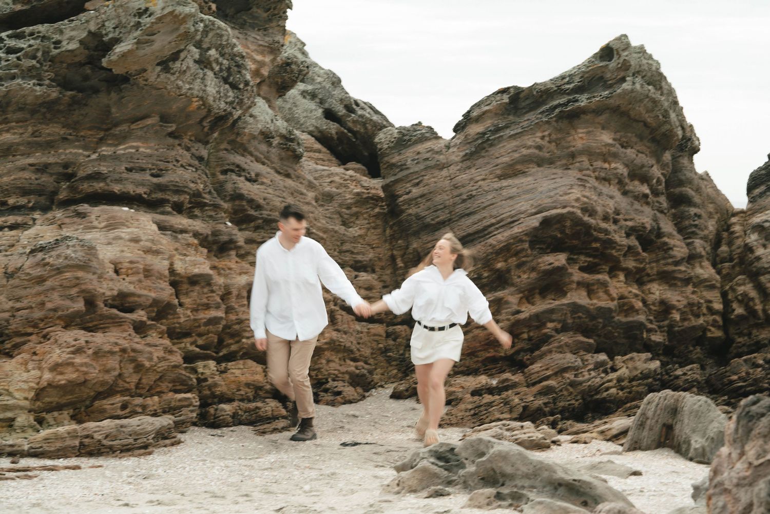 Romantic couple explores rocky coastal landscape at sunset among dramatic cliffs and ocean waves.