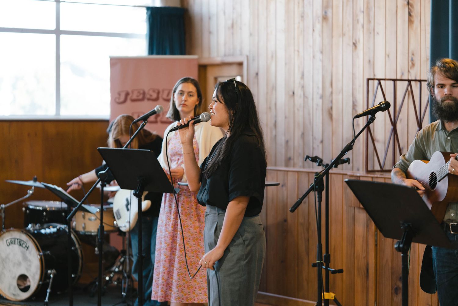 Musicians perform on stage with instruments and microphones in a wooden-paneled church setting.