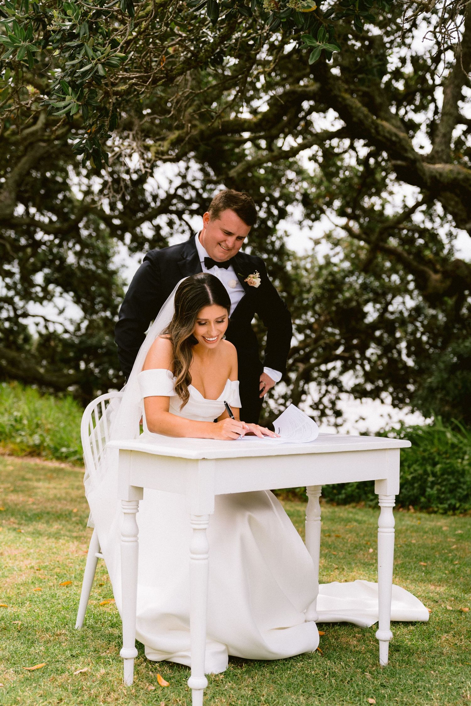 A couple in wedding attire sits at an elegant white desk on a grassy lawn surrounded by trees.