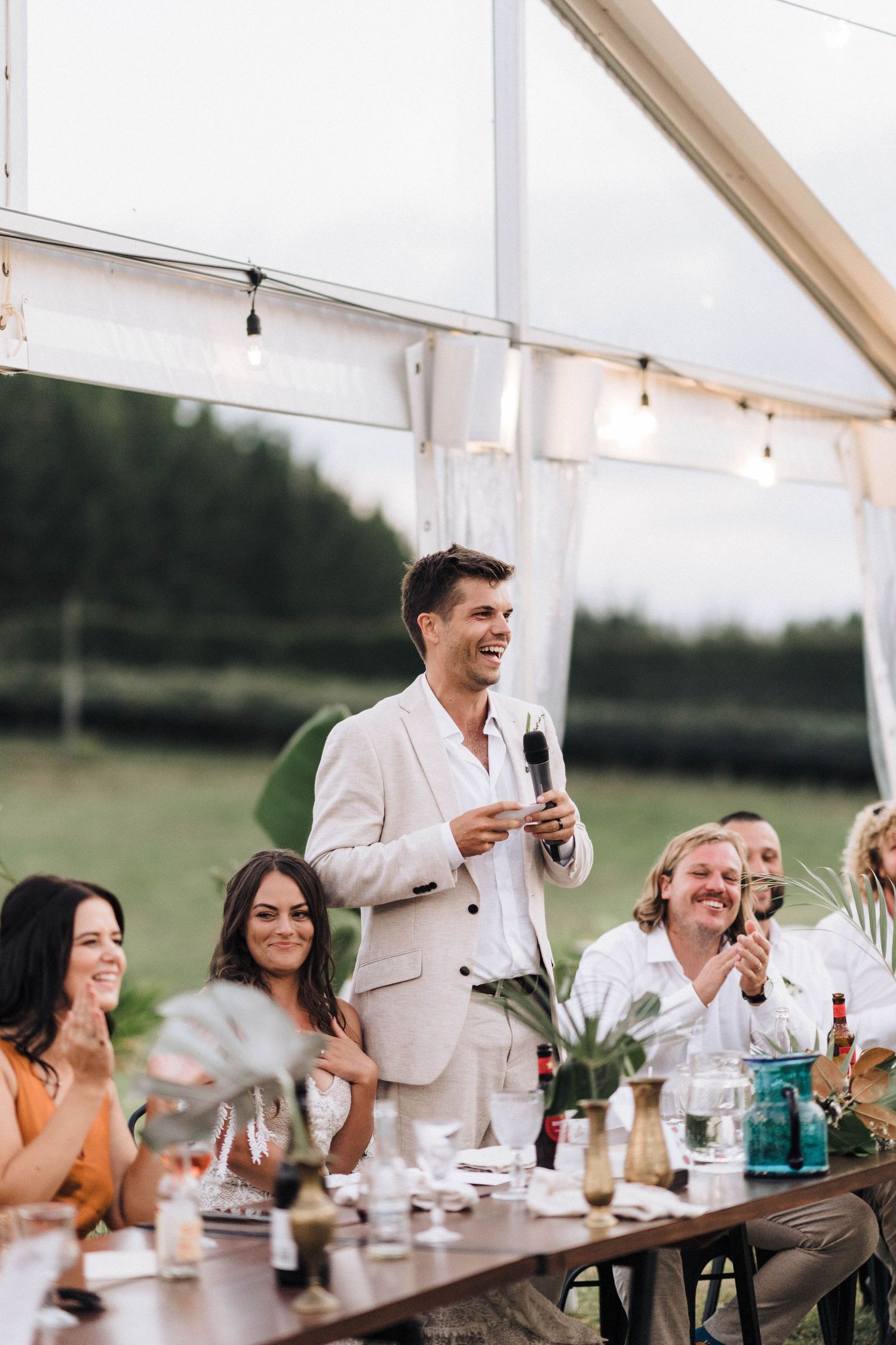 Wedding guest gives toast during outdoor reception under tent with string lights.