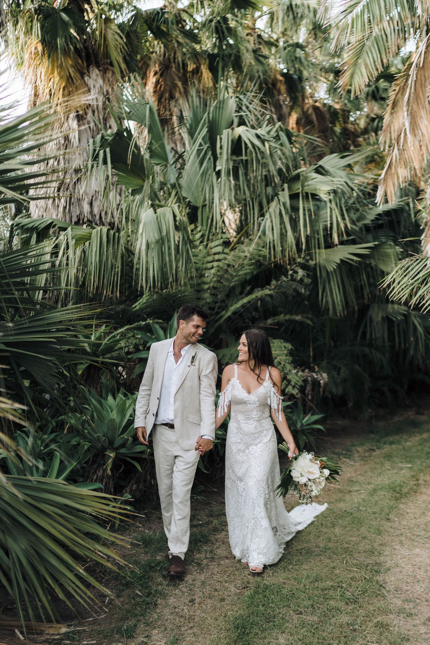 A tropical wedding scene with newlyweds walking along a path beneath lush palm trees and greenery.